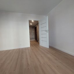 Jarbud - Jasne wnętrze po remoncie: białe ściany, panele podłogowe w odcieniach drewna i białe drzwi z poziomymi frezami. Widok na sąsiednie pomieszczenie z szarą ścianą.