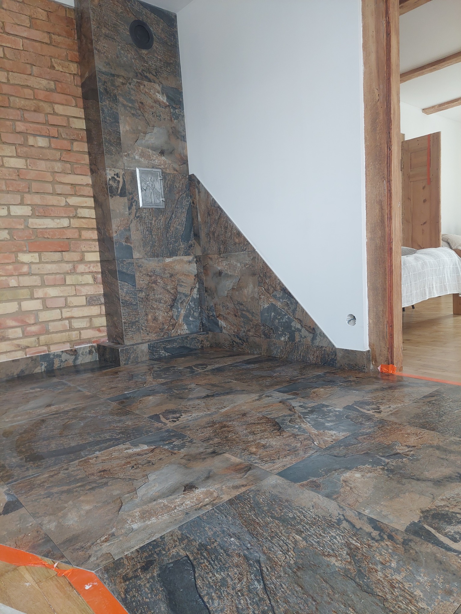 Wykończony fragment wnętrza z brązowo-szarymi płytkami imitującymi kamień na podłodze i ścianie, z widoczną ceglaną ścianą i drewnianą framugą drzwi.