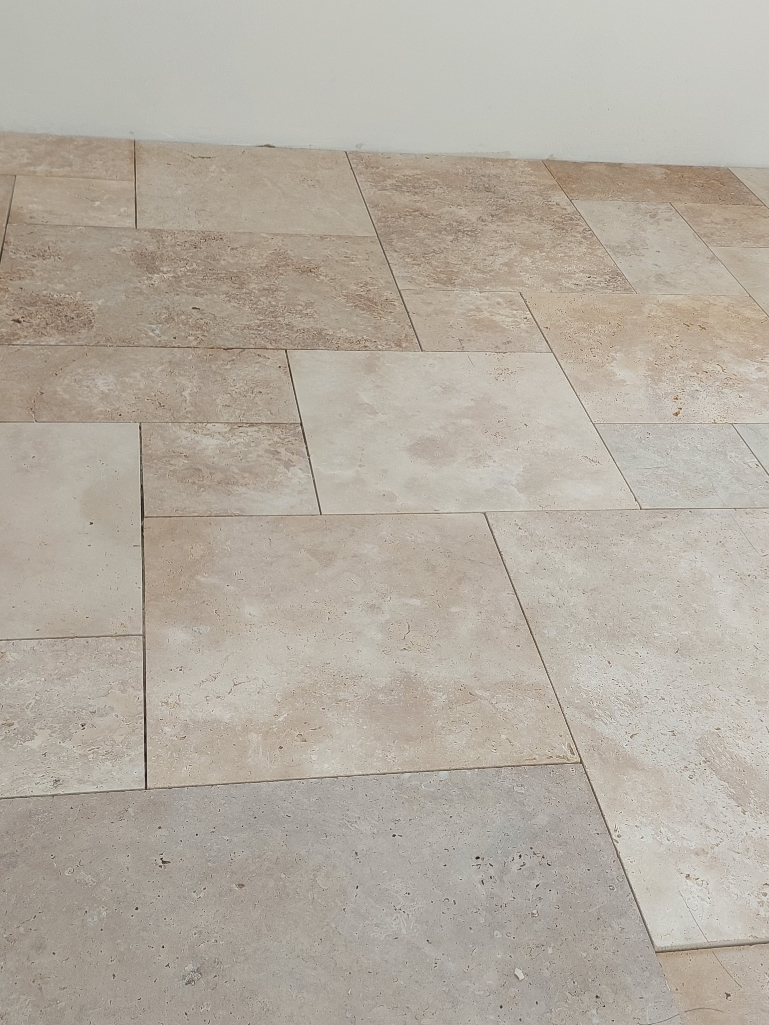 Beżowa podłoga z płytek o zróżnicowanych rozmiarach i fakturach, ułożonych w nieregularny wzór. Widoczne fugi i fragment białej ściany w tle.