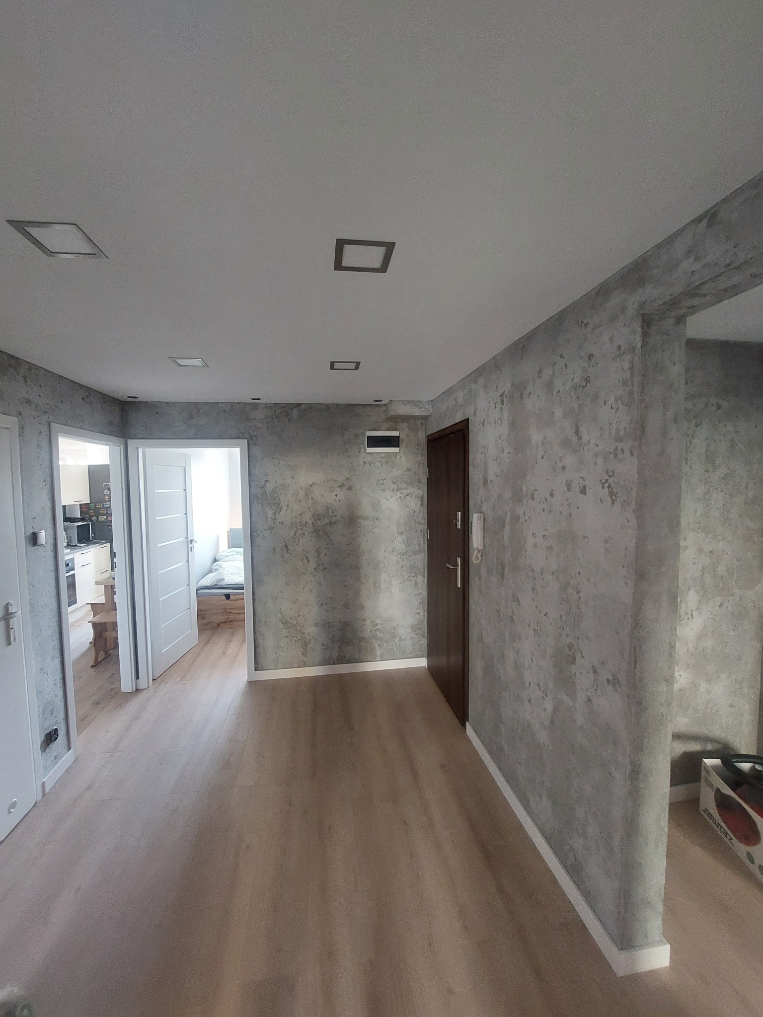 Wnętrze mieszkania po remoncie: szare ściany imitujące beton, drewnopodobna podłoga, białe drzwi i listwy przypodłogowe. Widoczne oświetlenie sufitowe.