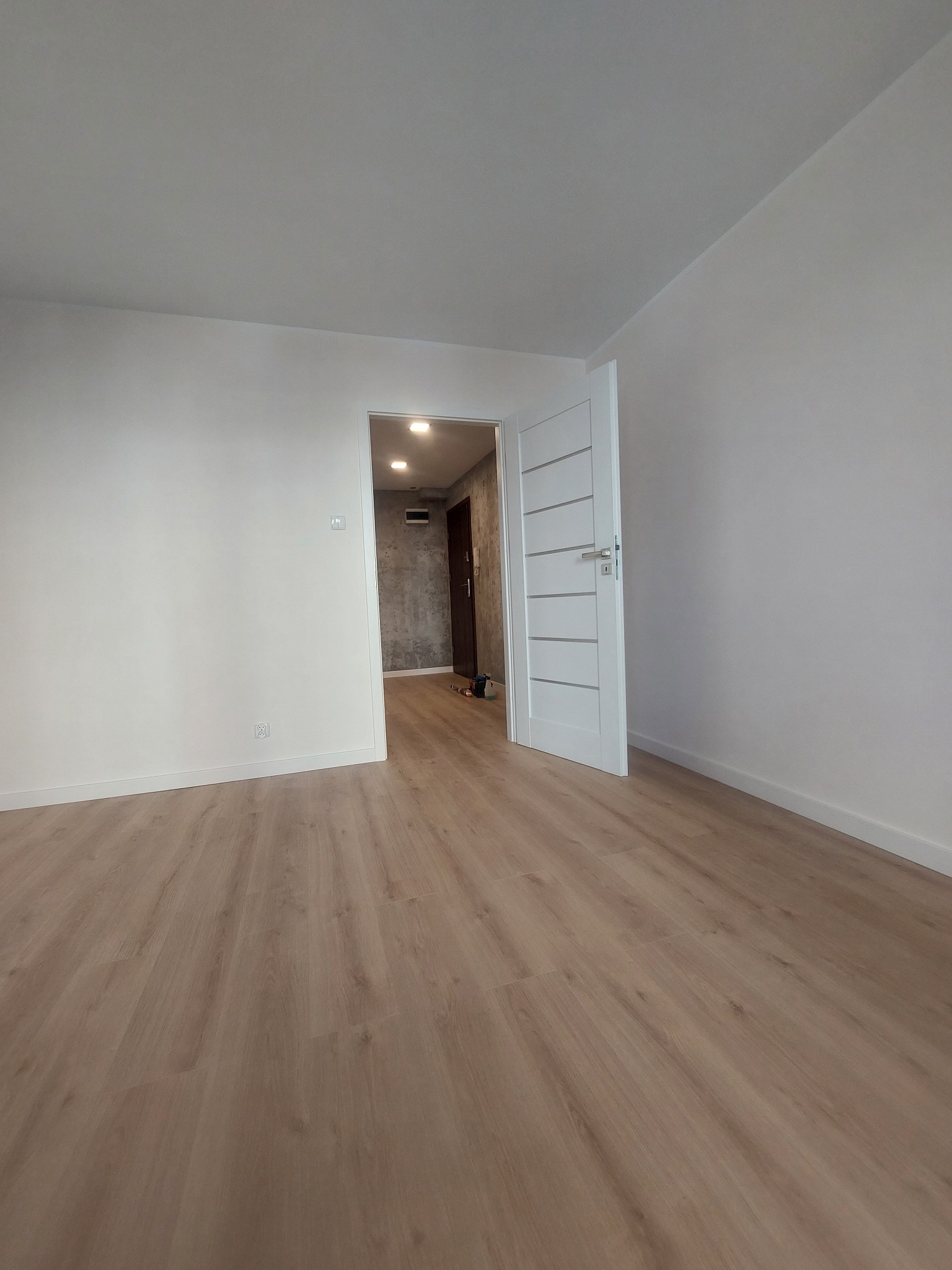 Jasne wnętrze po remoncie: białe ściany, panele podłogowe w odcieniach drewna i białe drzwi z poziomymi frezami. Widok na sąsiednie pomieszczenie z szarą ścianą.