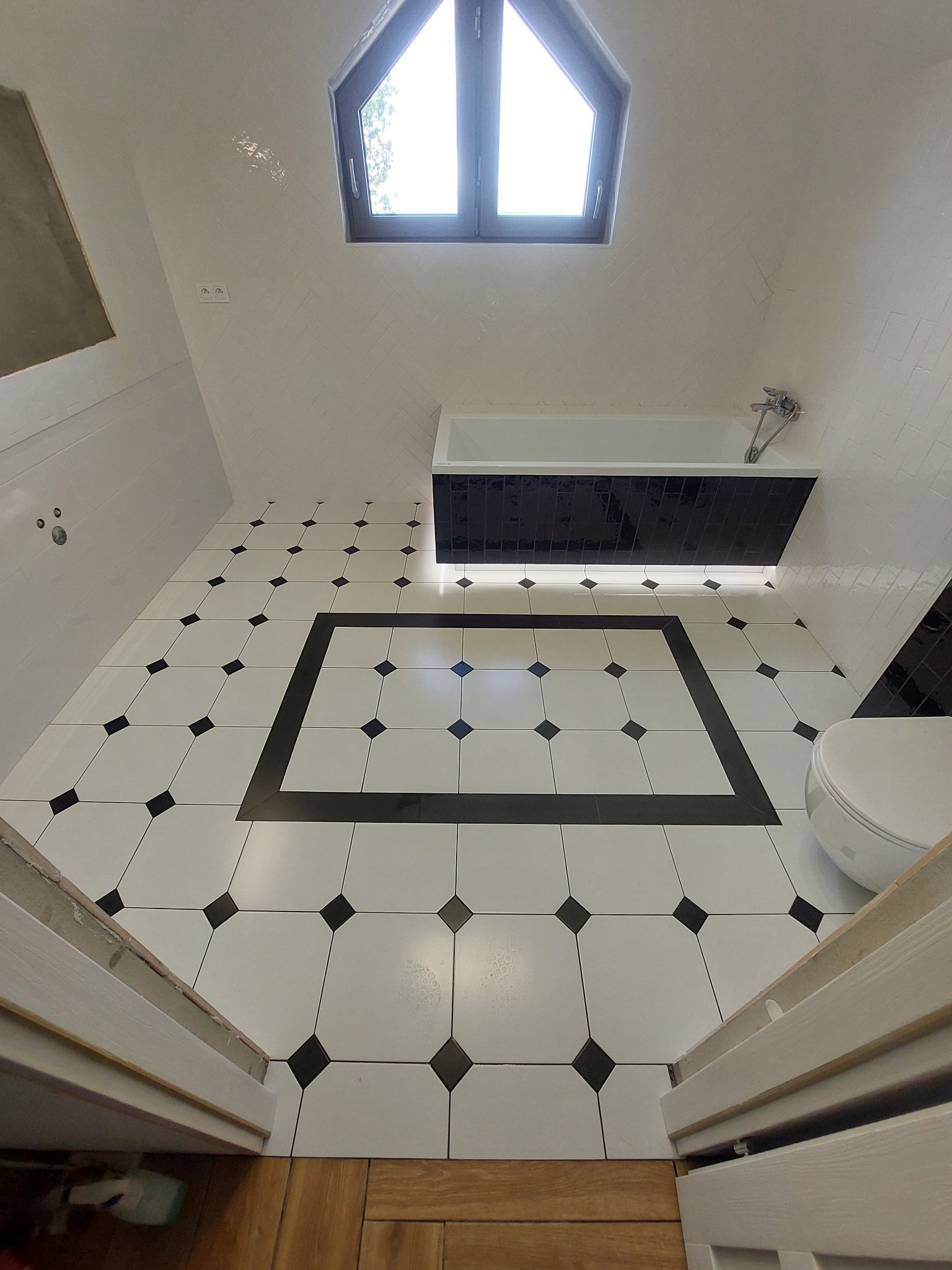 Biała łazienka z czarnymi akcentami: wzorzysta podłoga, obudowa wanny i okno dachowe. Widok z góry, drzwi wejściowe z drewna w dolnej części kadru.