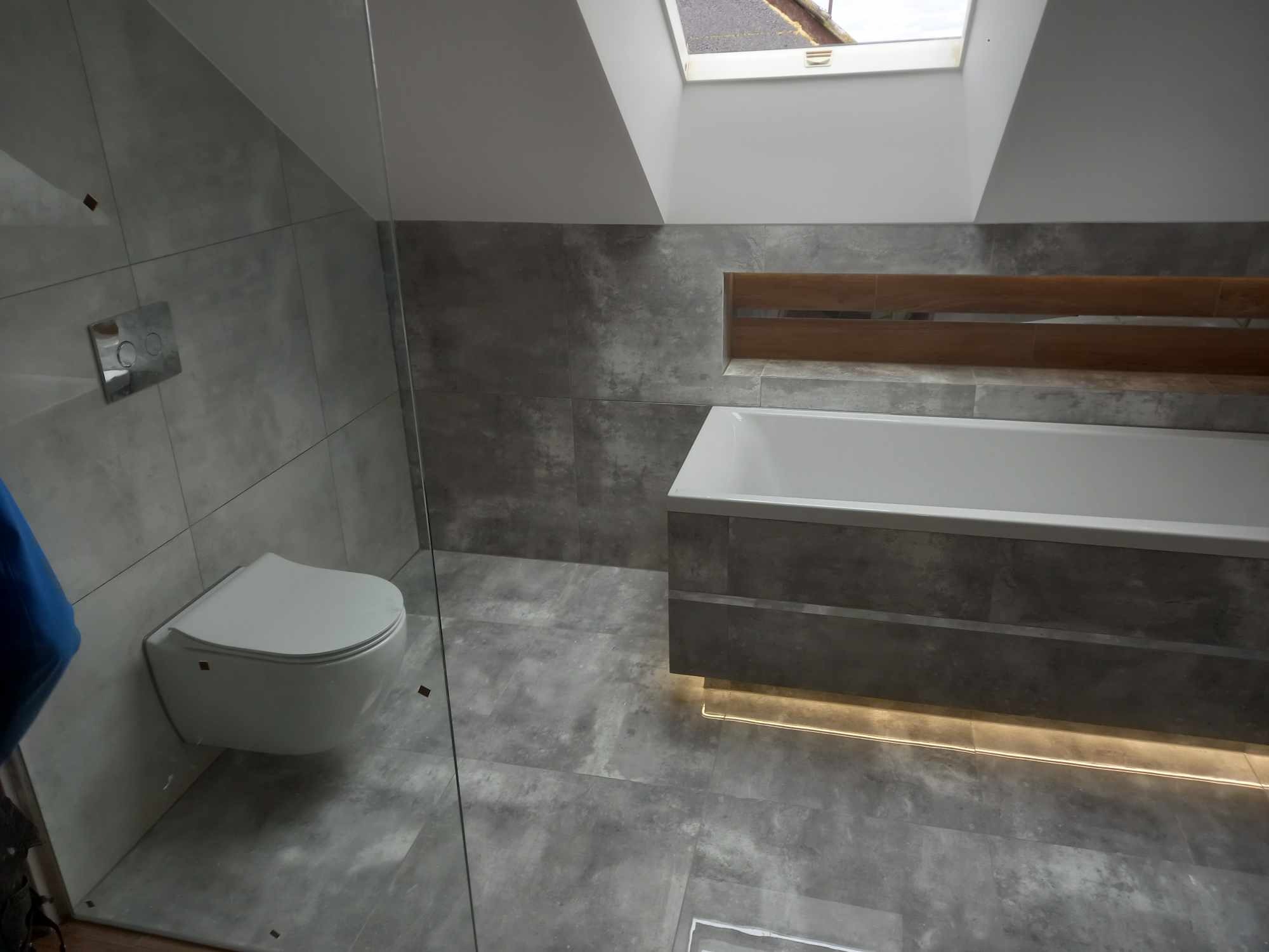 Nowoczesna łazienka w szarościach z podwieszaną toaletą, oświetleniem LED pod wanną i oknem dachowym. Minimalistyczny design z akcentami drewna.