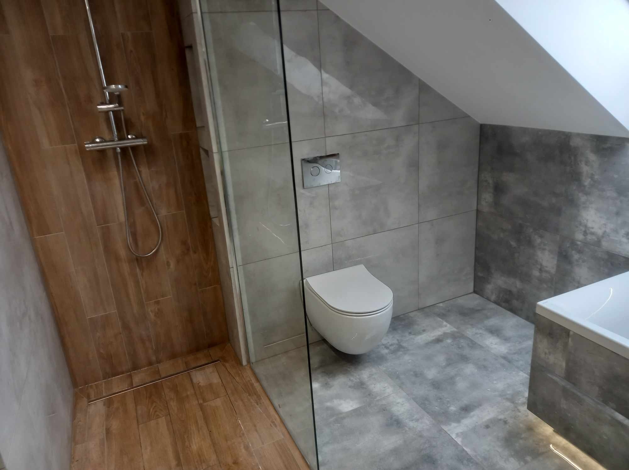 Nowoczesna łazienka na poddaszu z kabiną prysznicową oddzieloną szklaną ścianką, wiszącą toaletą i szarą glazurą. Widoczny odpływ liniowy.