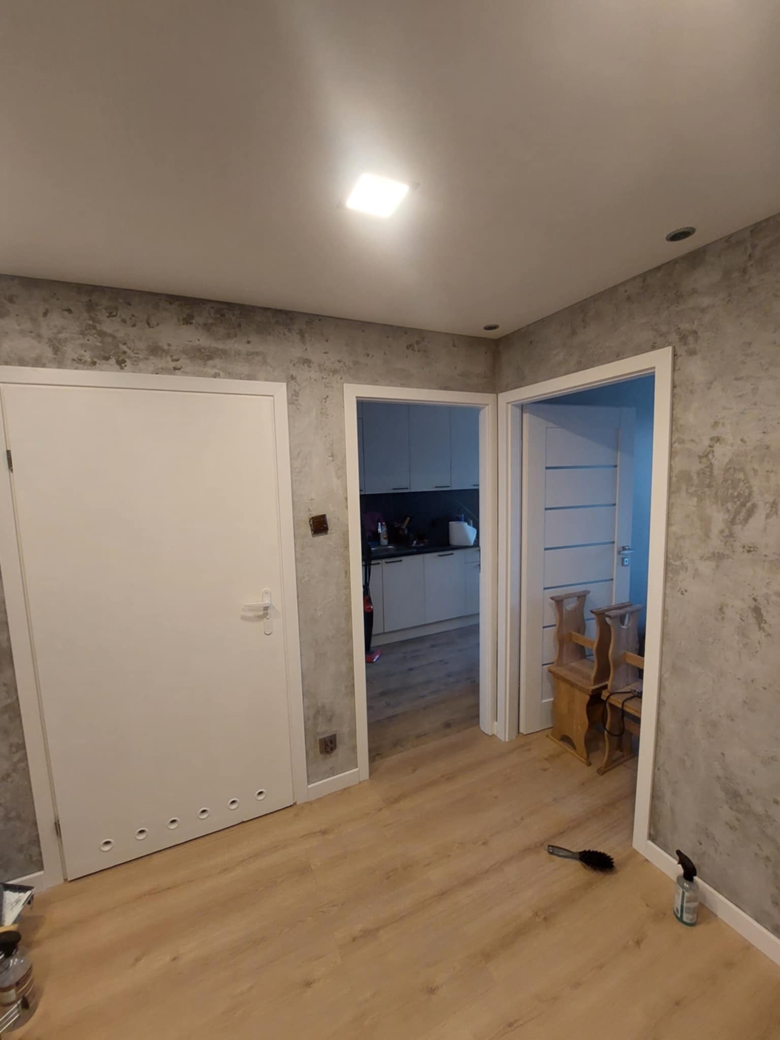 Wykończone wnętrze: szara ściana z efektem betonu, białe drzwi z otworami wentylacyjnymi u dołu, drewniana podłoga i widok na kuchnię w tle.