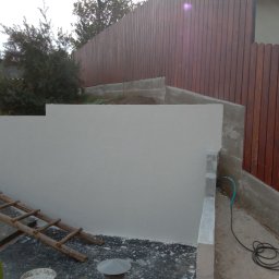 Brak - Świeżo otynkowana, jasna ściana oporowa z drewnianym płotem w tle. Widoczna drabina, narzędzia i pozostałości po tynkowaniu. Ujęcie z dołu, podkreślające strukturę tynku.