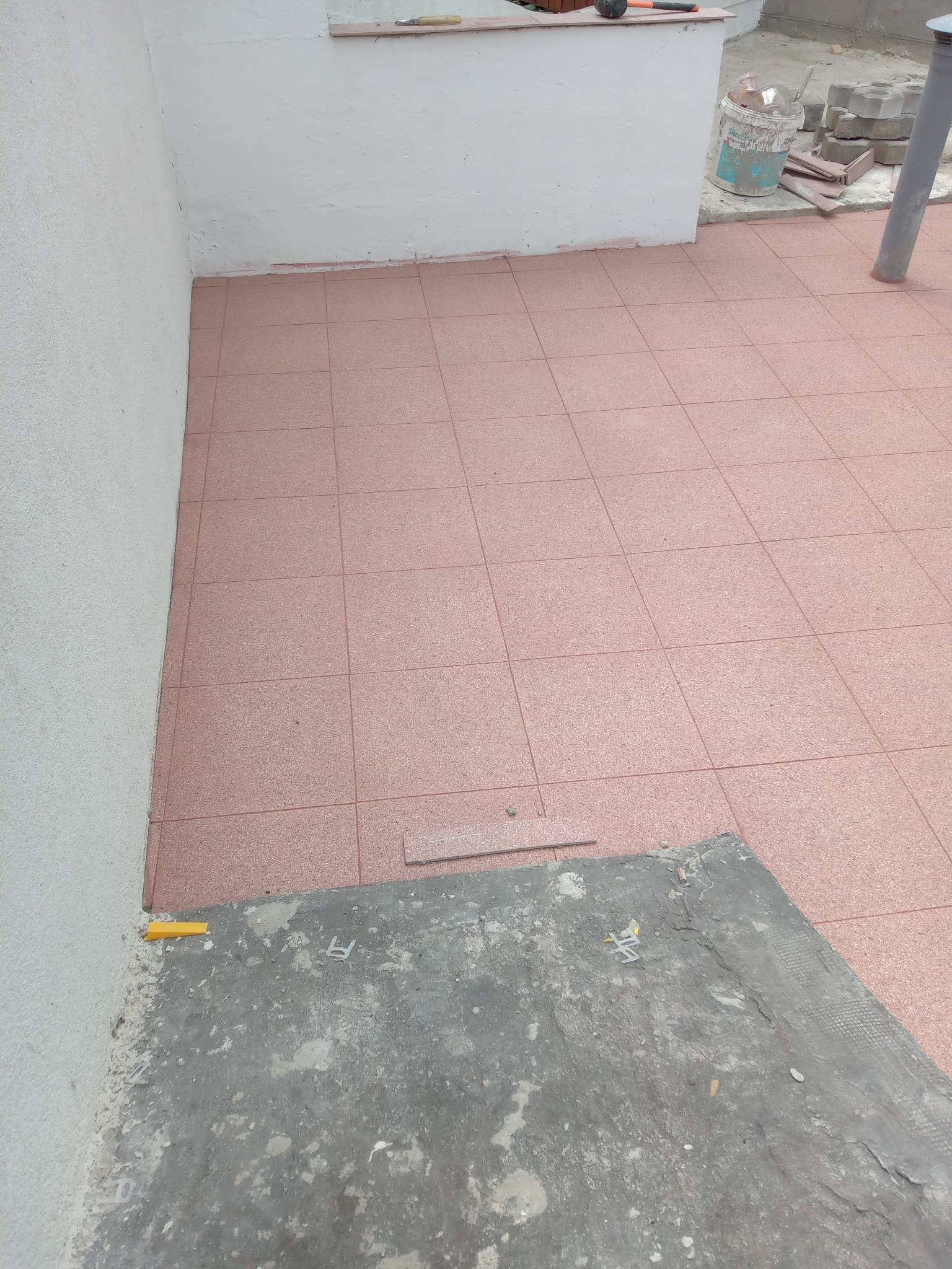 Ułożona, różowa terakota na balkonie, widoczne narzędzia glazurnicze i fragment ściany. Częściowo ukończone prace wykończeniowe na zewnątrz budynku.