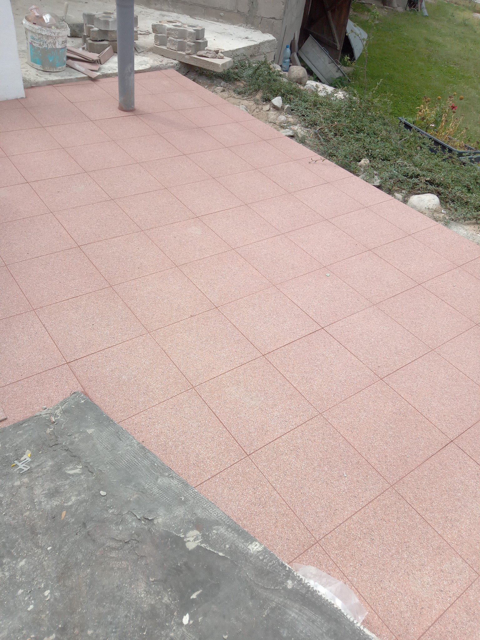 Ułożona, różowa kostka brukowa na zewnątrz budynku, z widocznymi elementami zieleni i narzędziami budowlanymi w tle. Częściowo ukończona nawierzchnia.