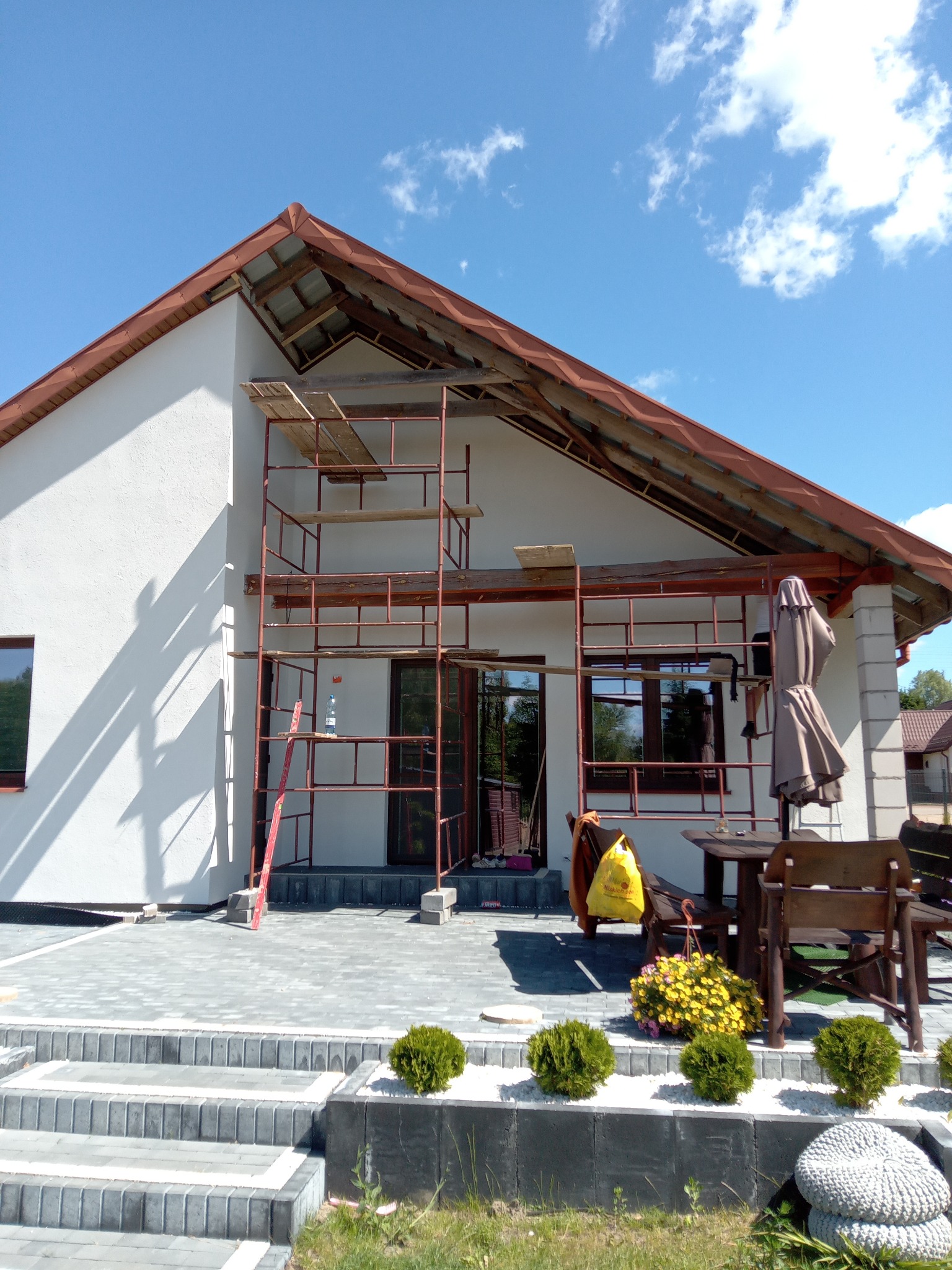 Dom jednorodzinny w trakcie prac elewacyjnych: biała ściana, brązowe rusztowanie, widoczne okna i drzwi. Uwagę przykuwa starannie wykonany taras z kostki brukowej.