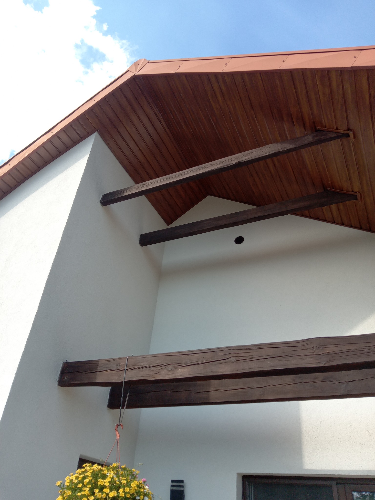 Biała elewacja domu z drewnianymi belkami pod brązowym podbitką dachową. W tle błękitne niebo. Wiszące kwiaty w rogu kadru.