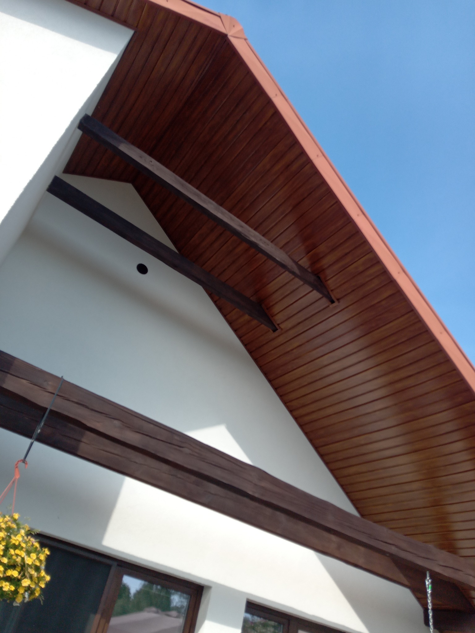 Biała elewacja domu z drewnianą podbitką i belkami, widok z dołu pod kątem, fragment okna i wiszące kwiaty w kadrze, błękitne niebo w tle.