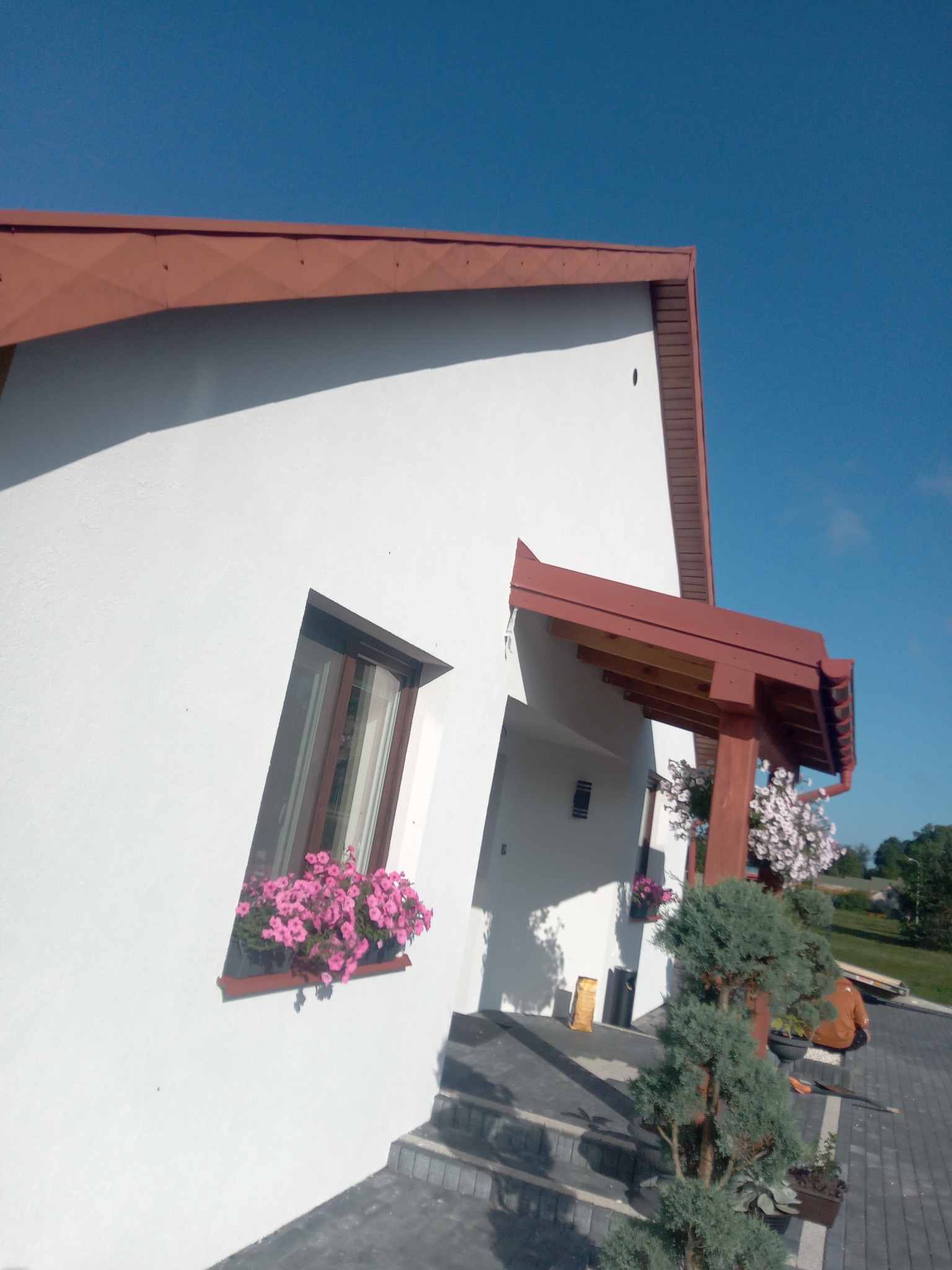 Biała elewacja domu z brązowym dachem i oknami, ozdobiona różowymi kwiatami w donicach. Widoczne schody wejściowe i fragment ogrodu. Ujęcie z dołu, błękitne niebo w tle.