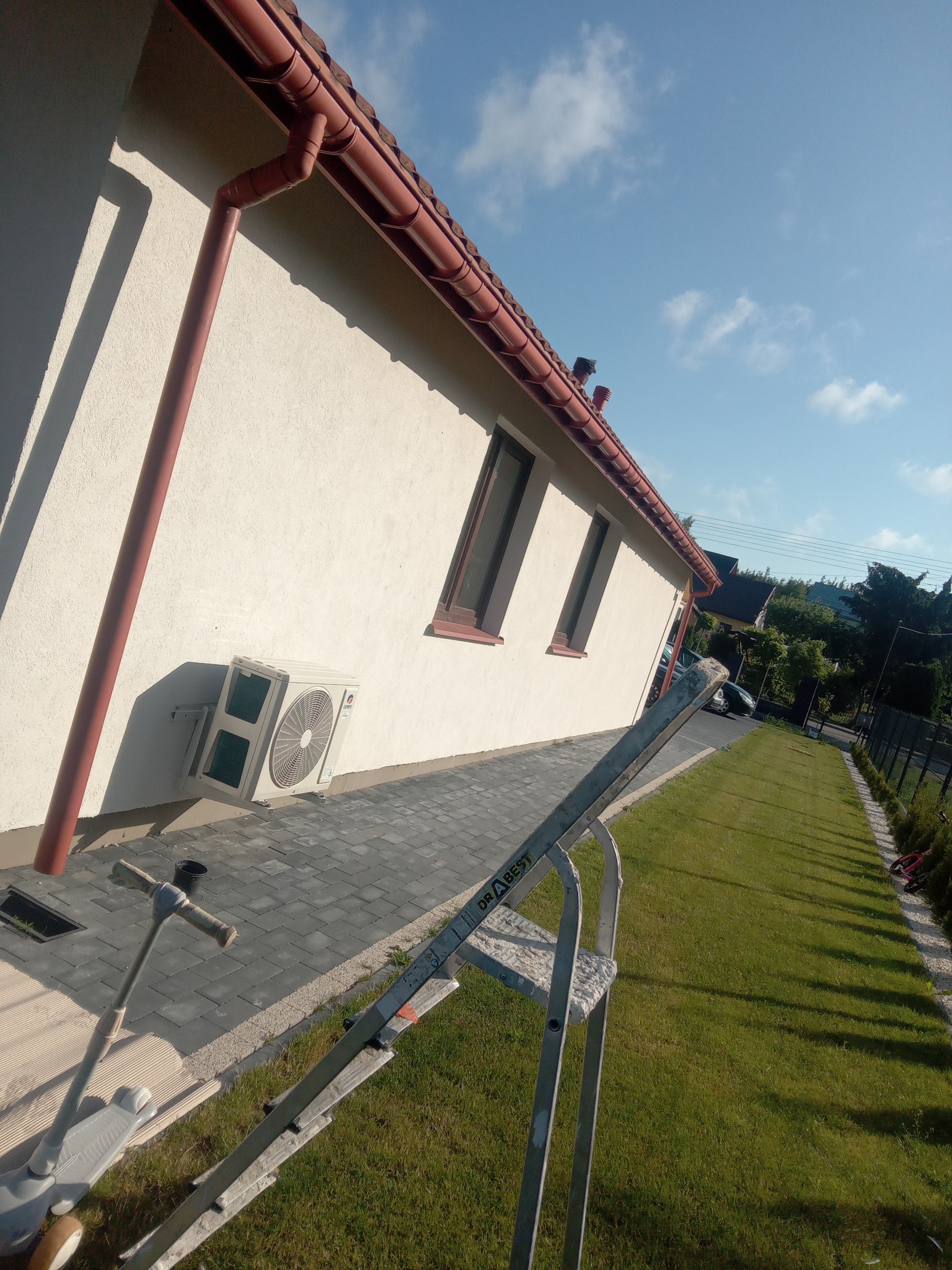 Jasna elewacja domu z brązowymi rynnami i oknami, widoczny klimatyzator na ścianie, trawnik i drabina oparta o ścianę. Ujęcie z dołu, pochmurne niebo.