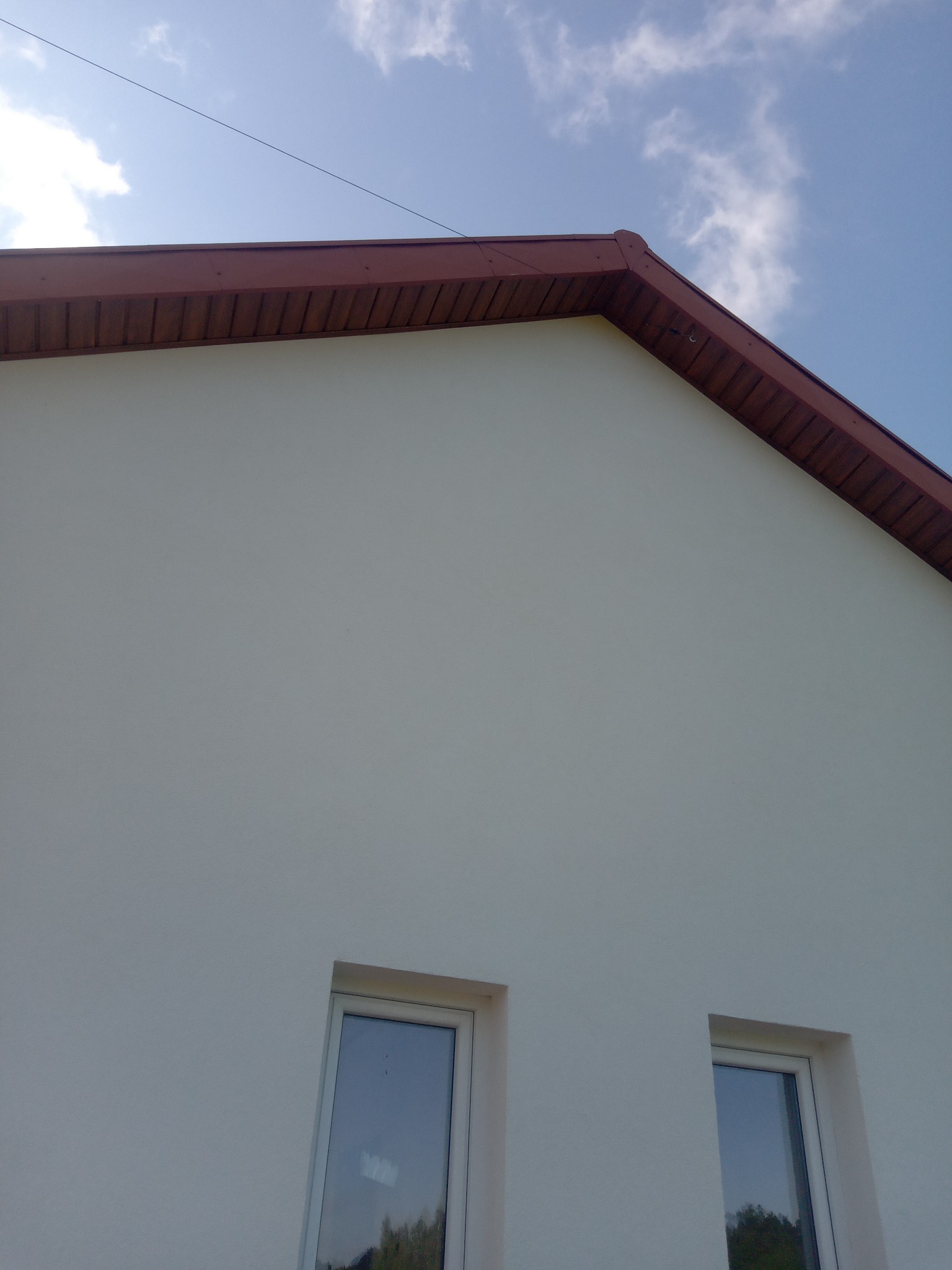 Biała elewacja domu jednorodzinnego z brązowym wykończeniem dachu i dwoma oknami, Sokółka. Widok z dołu na fragment ściany i niebo.