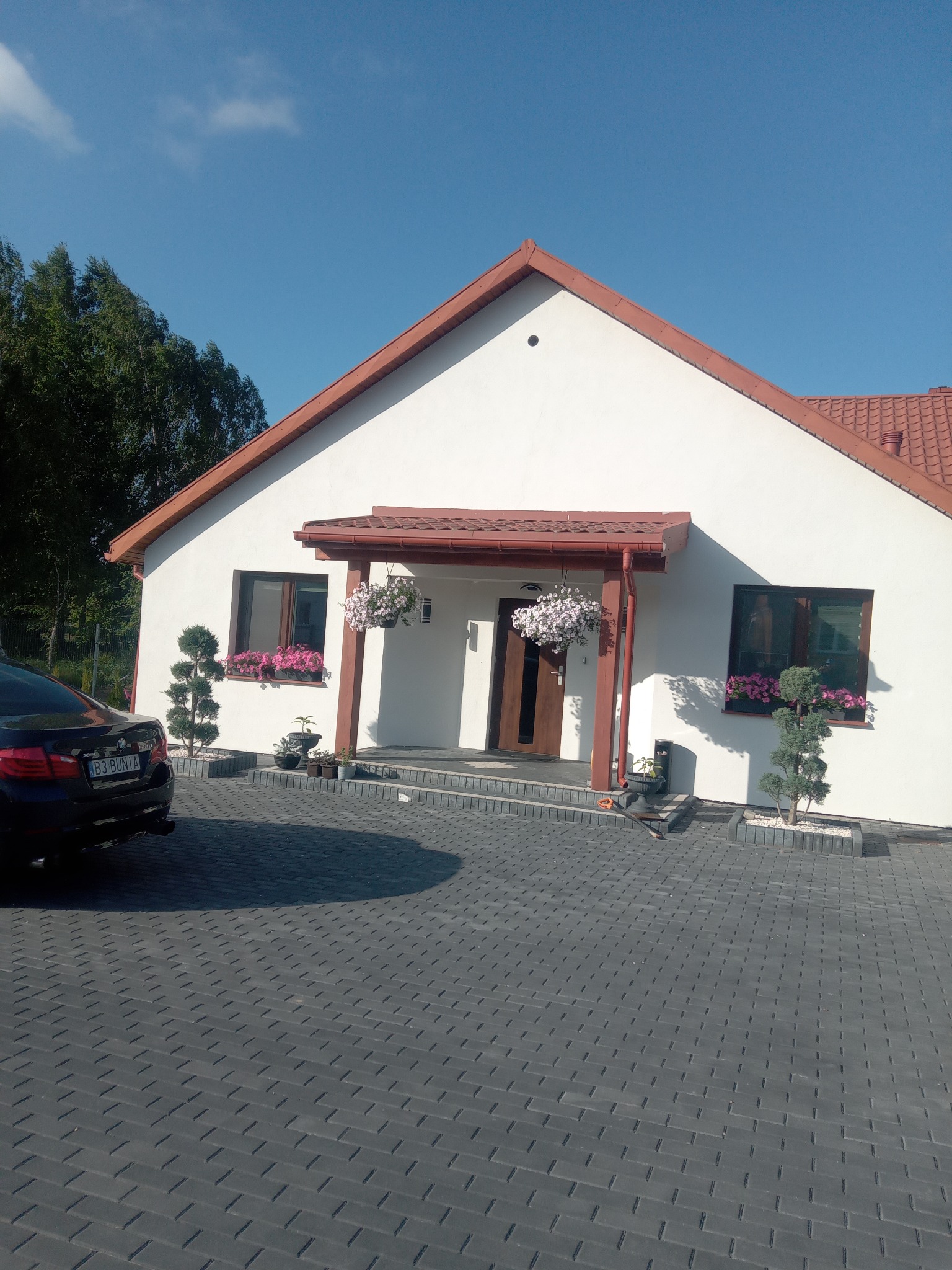 Biały dom z czerwoną dachówką i zadbanym otoczeniem: kostka brukowa, kwiaty w oknach i wiszące donice. Widoczny fragment czarnego samochodu.