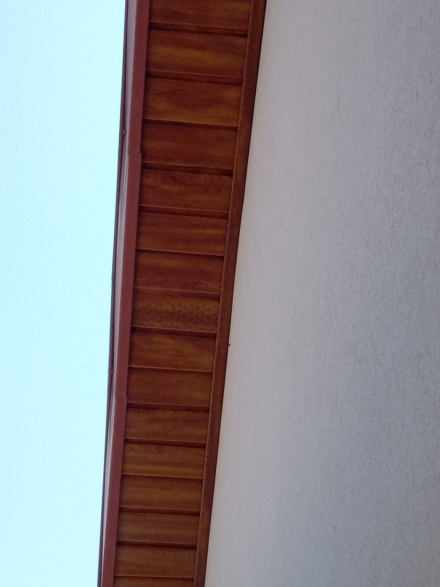 Fragment elewacji budynku z drewnianą podbitką w ciepłym odcieniu brązu, kontrastujący z jasną, teksturowaną ścianą i błękitnym niebem w tle.