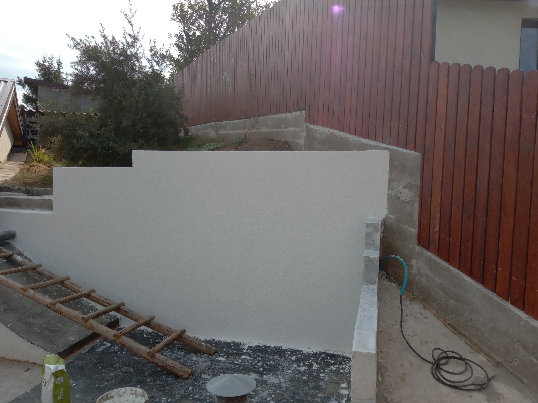 Świeżo otynkowana, jasna ściana oporowa z drewnianym płotem w tle. Widoczna drabina, narzędzia i pozostałości po tynkowaniu. Ujęcie z dołu, podkreślające strukturę tynku.