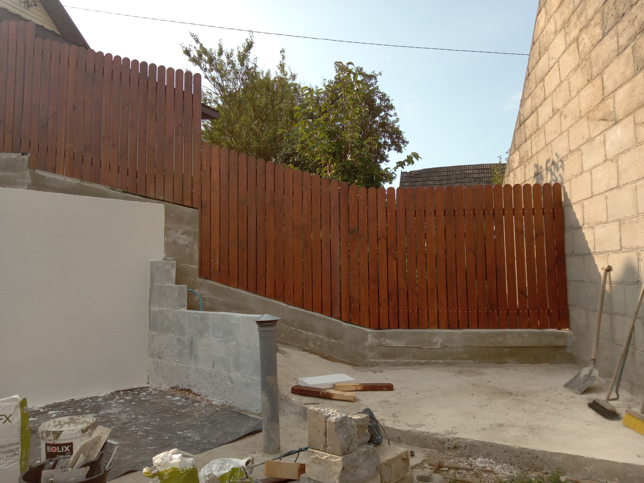 Elewacja domu w trakcie prac wykończeniowych: otynkowana ściana, drewniany płot w tle, narzędzia i materiały budowlane na ziemi. Widoczne worki z zaprawą.