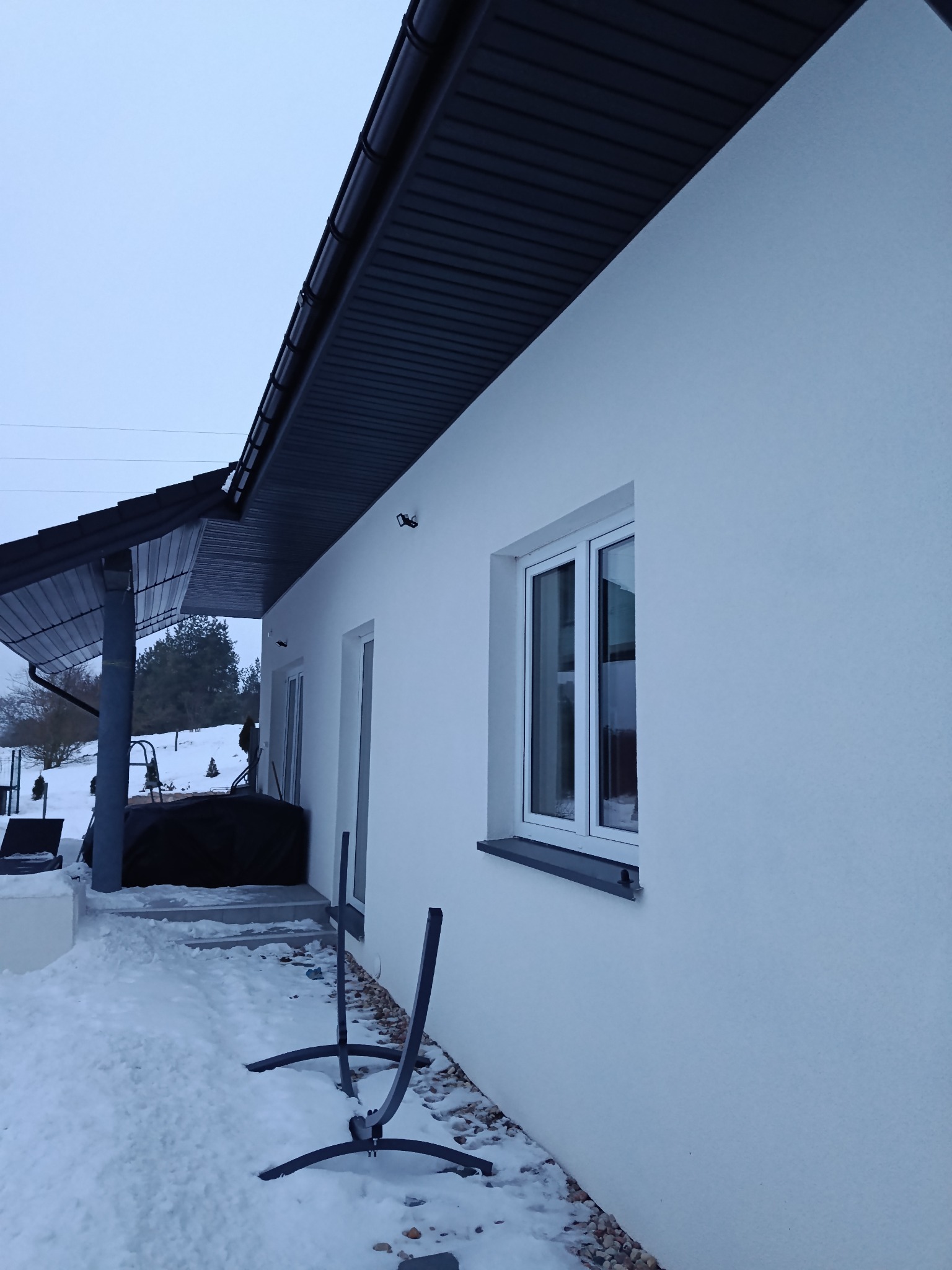 Biała elewacja domu jednorodzinnego w zimowej scenerii. Widoczne okno z szarą ramą, fragment dachu i zasypany śniegiem teren wokół budynku. Minimalistyczny design.