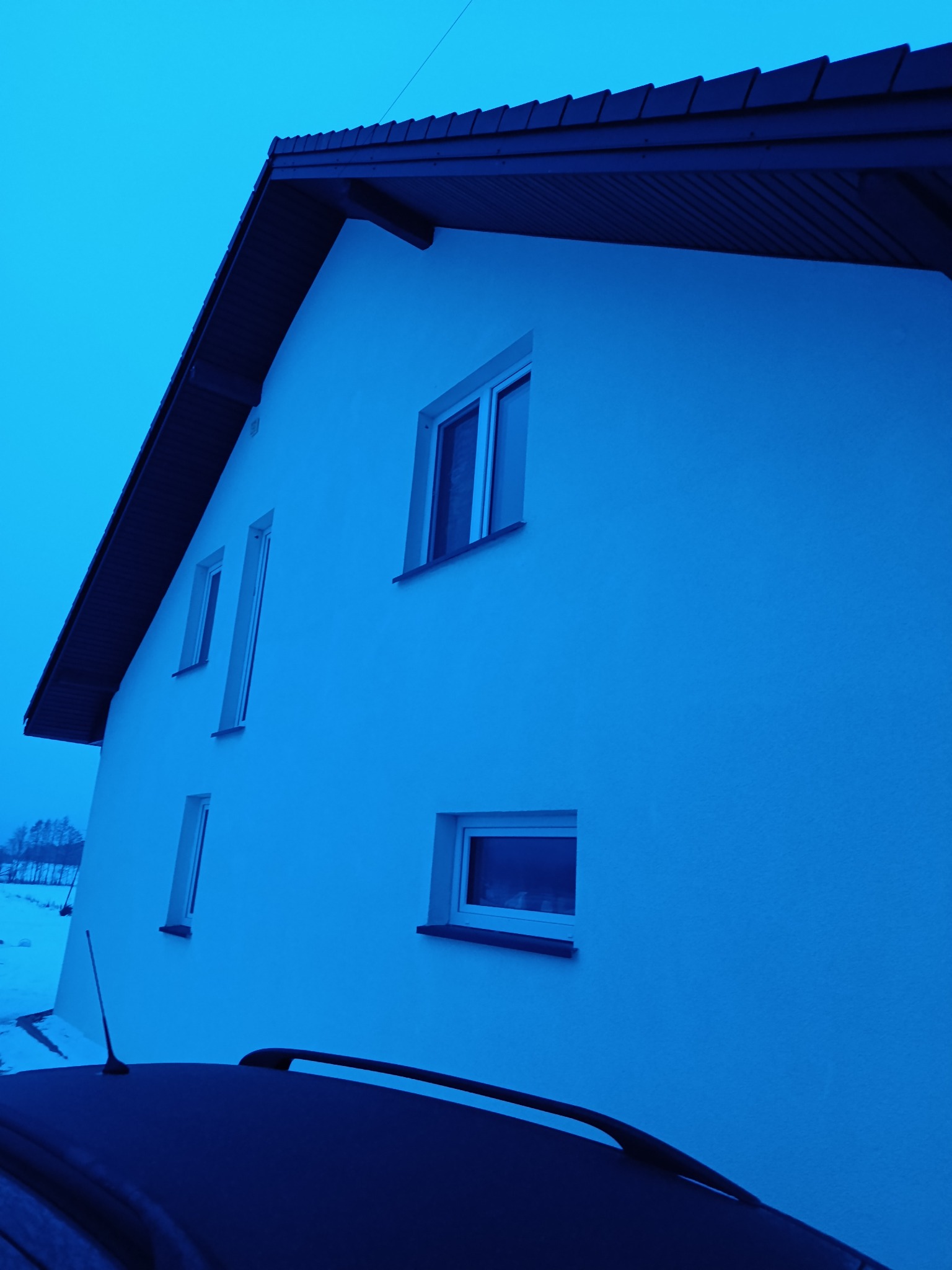 Boczny widok na biały dom z ciemnym dachem i kilkoma oknami, sfotografowany z dołu, z fragmentem dachu samochodu w kadrze. Zimowa sceneria w tle.