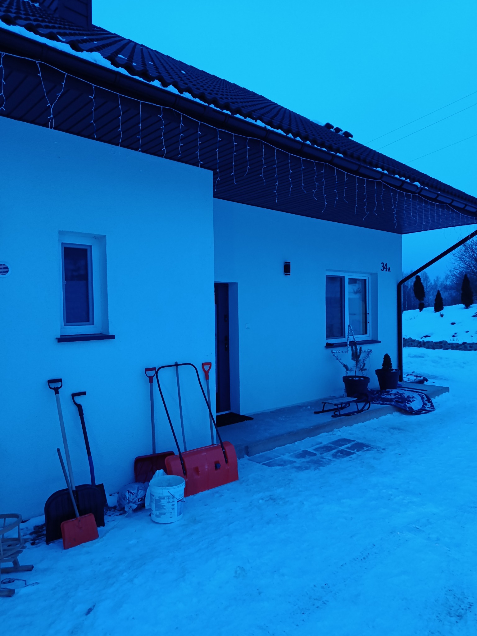 Biały dom jednorodzinny z numerem 34A, oświetlony świątecznymi lampkami, z narzędziami do odśnieżania opartymi o ścianę. Zima, śnieg.