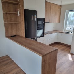 IDEA Damian Dyoniziak - Nowoczesna kuchnia z drewnianymi akcentami: białe szafki, drewniany blat i półki, czarna lodówka. Widoczne oświetlenie w postaci żarówki na kablu.