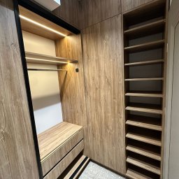 IDEA Damian Dyoniziak - Funkcjonalna szafa wnękowa z przesuwnymi drzwiami i oświetleniem LED, wykończona w ciepłym odcieniu drewna. Widoczne półki i szuflady z czarnymi akcentami.