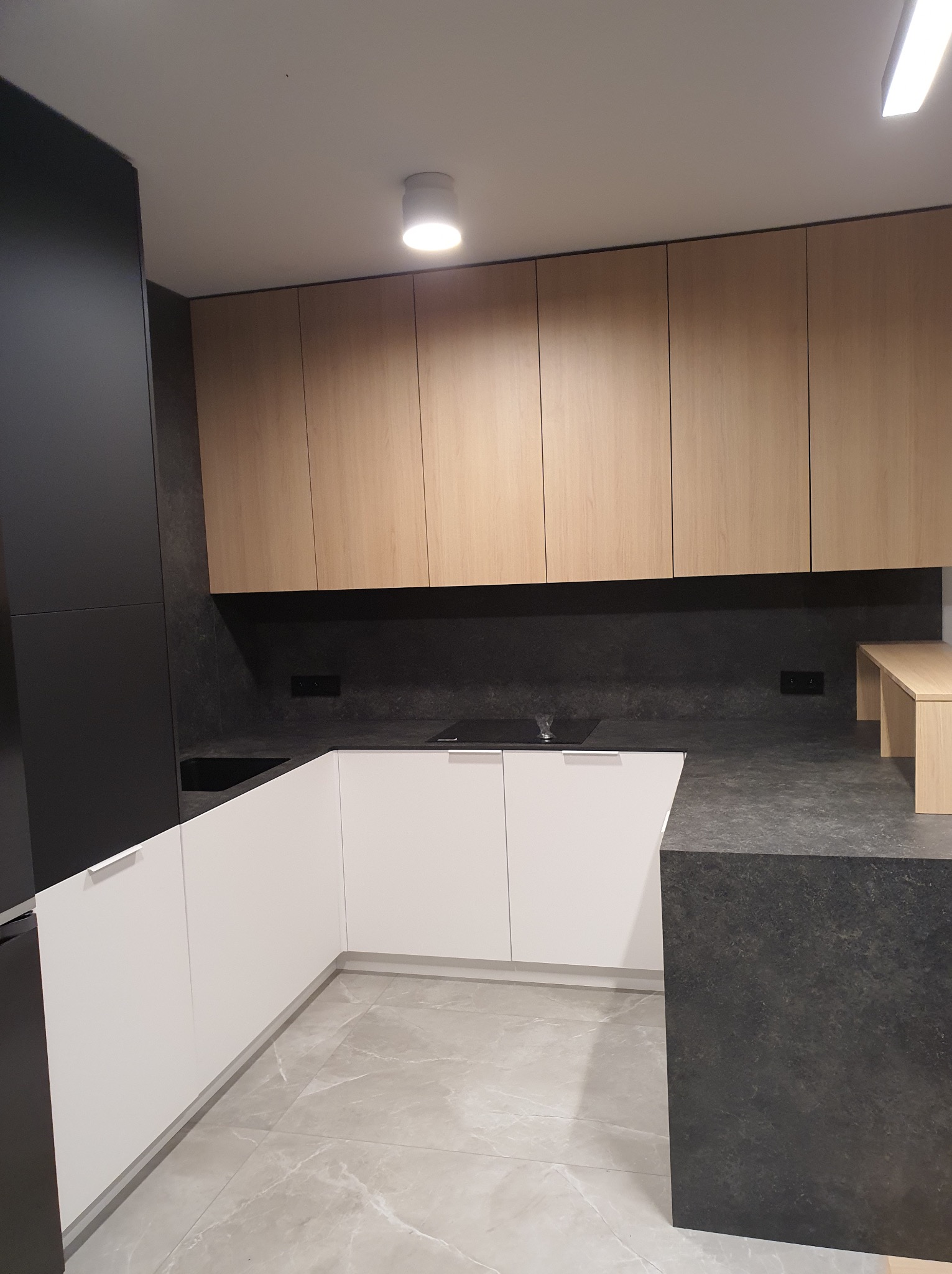 Nowoczesna kuchnia: białe dolne szafki, drewniane górne, czarny blat i ściana. Minimalistyczny design, szare płytki podłogowe. Oświetlenie sufitowe.