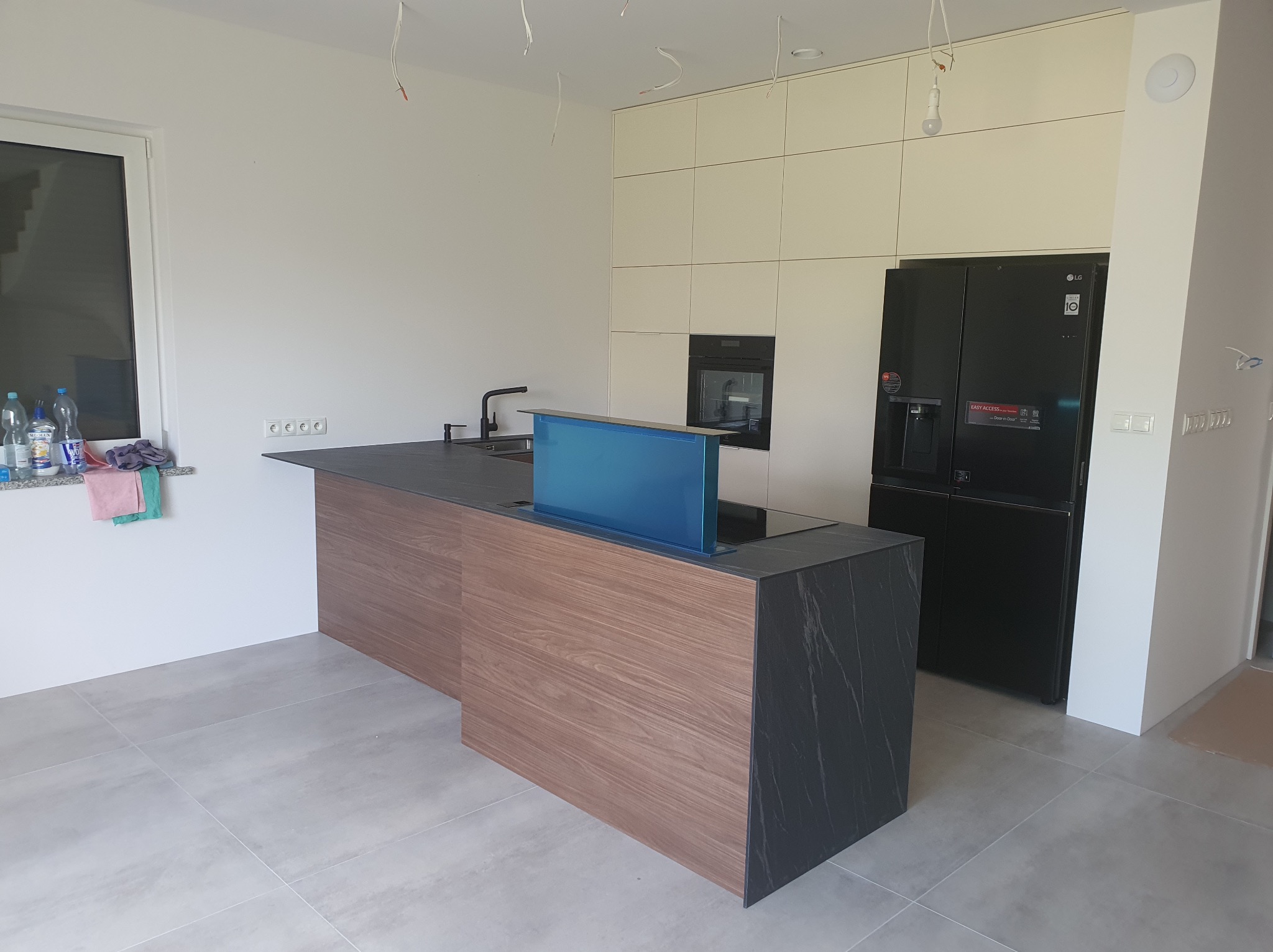 Nowoczesna kuchnia z wyspą: drewniany front, czarny blat, wysuwany okap. Białe szafki wiszące i czarna lodówka. Minimalistyczny design, surowe wykończenie.