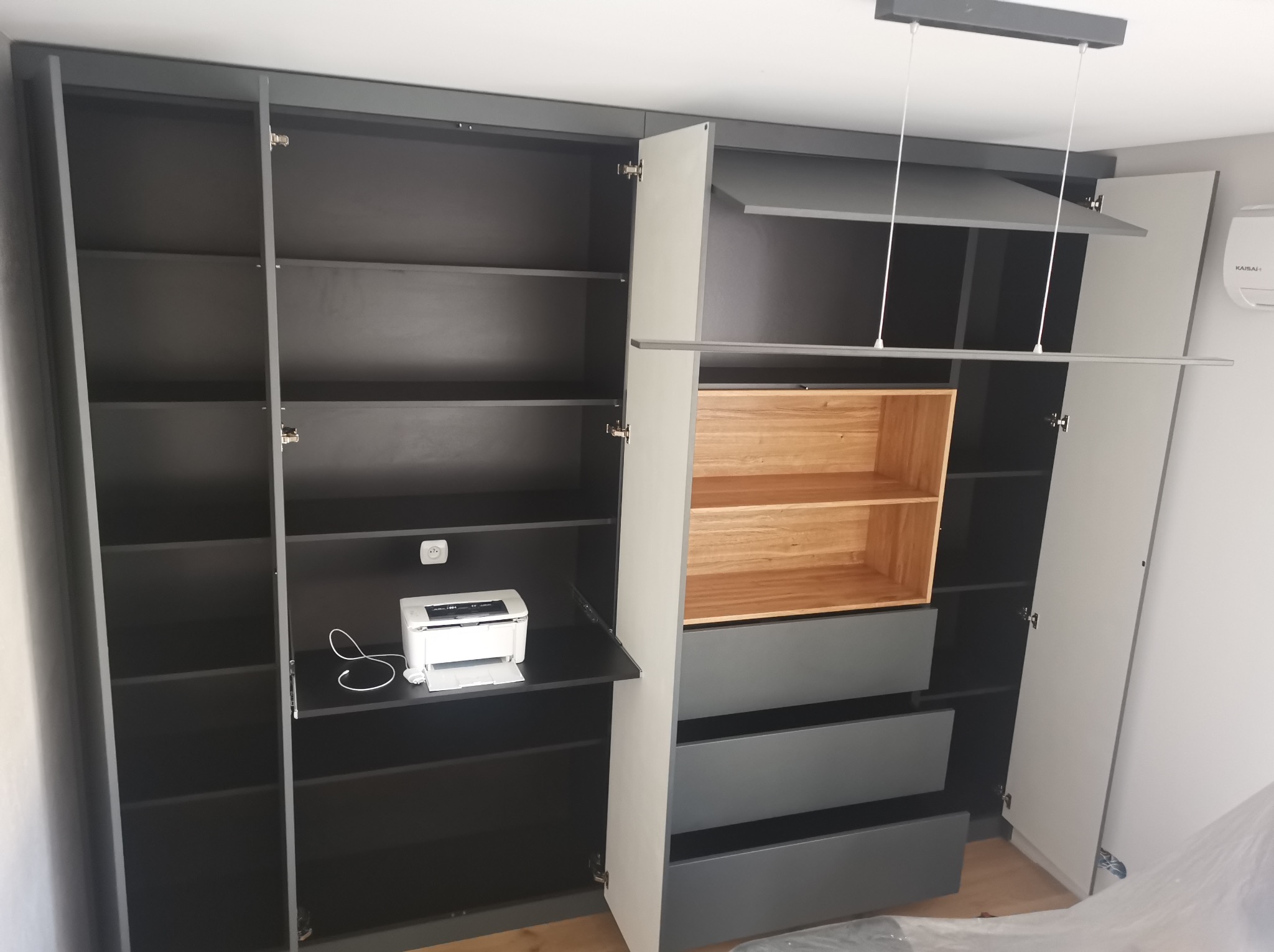 Nowoczesna szafa wnękowa z szarymi frontami, drewnianymi półkami i wbudowanym miejscem na drukarkę. Otwarta przestrzeń z półkami i szufladami.