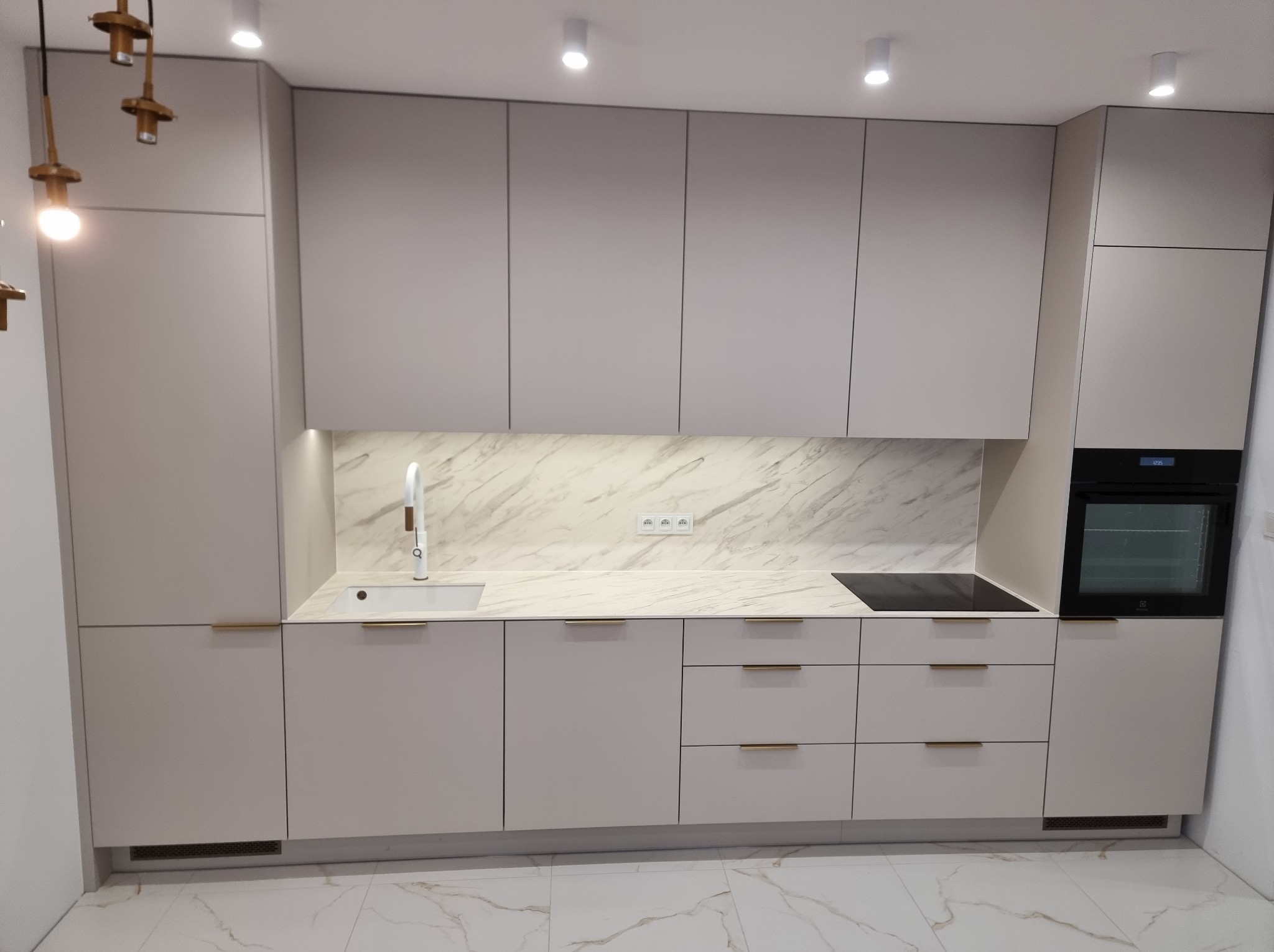 Jasna kuchnia z marmurowym blatem i złotymi uchwytami, oświetlona punktowymi lampami sufitowymi. Minimalistyczny design, nowoczesne wykończenie.