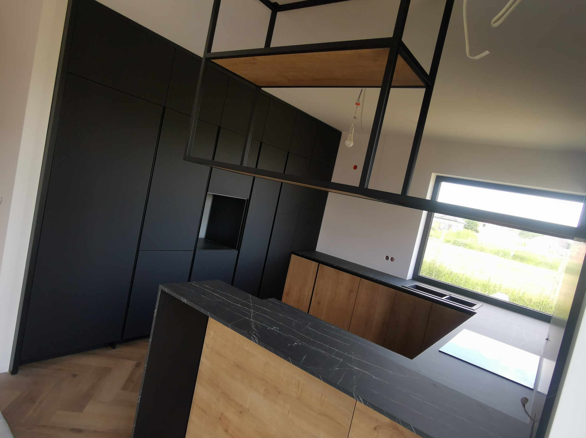 Nowoczesna kuchnia z czarnymi szafkami i drewnianym blatem, oświetlona naturalnym światłem z okna. Widoczny fragment wyspy kuchennej i metalowa konstrukcja nad nią.