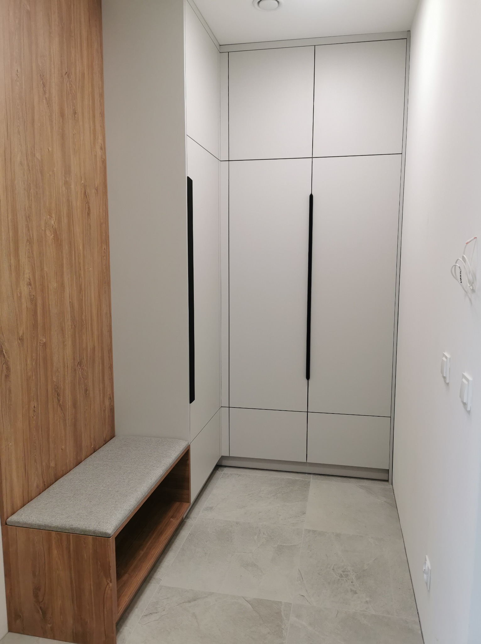 Nowoczesna szafa wnękowa z siedziskiem w przedpokoju. Białe fronty szafy z długimi, czarnymi uchwytami. Drewniane wykończenie siedziska i panelu ściennego.