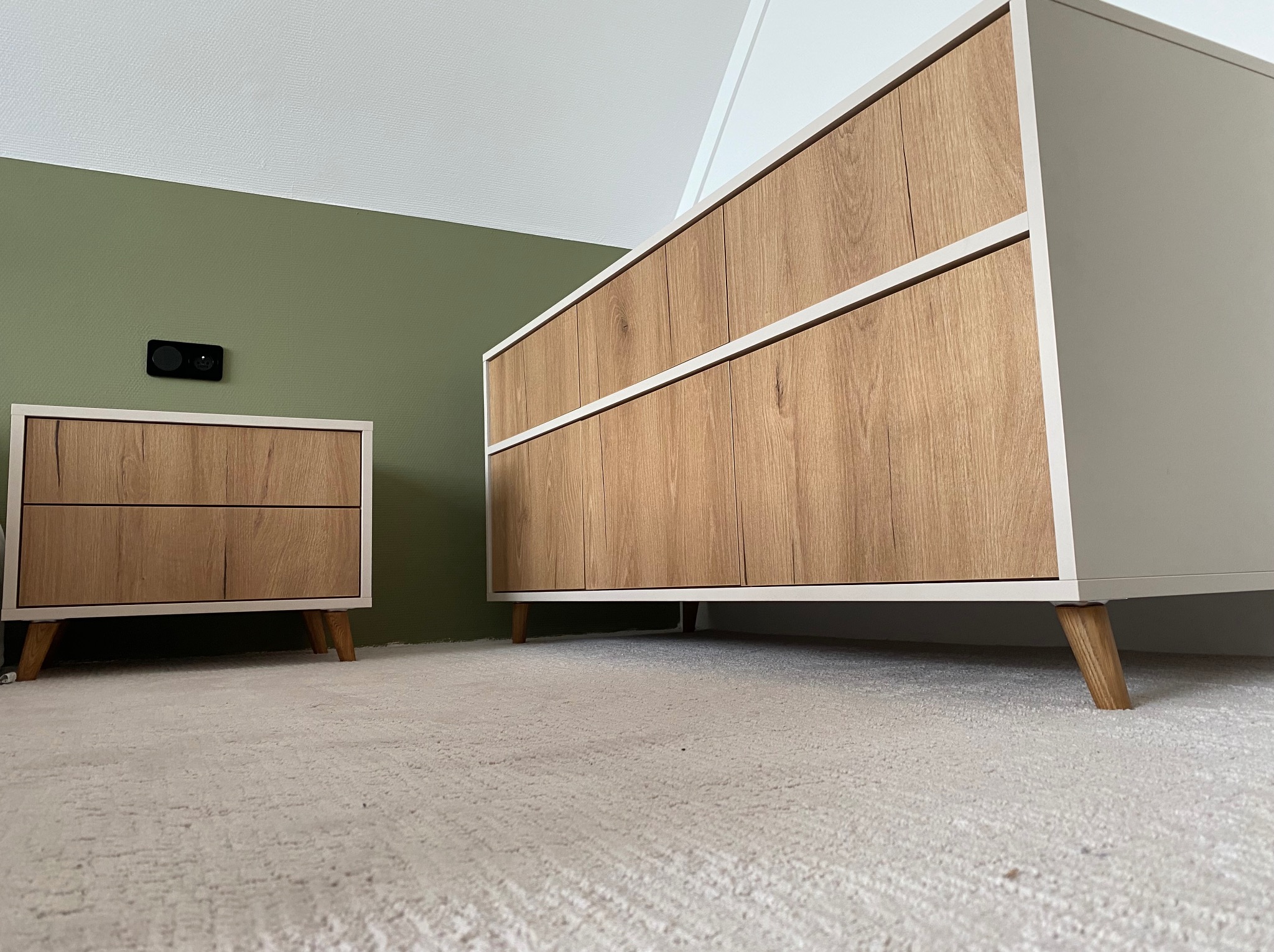 Komplet mebli: szafka nocna i komoda z drewnianymi frontami i jasnymi korpusami na drewnianych nóżkach, na tle zielonej ściany i jasnej wykładziny.