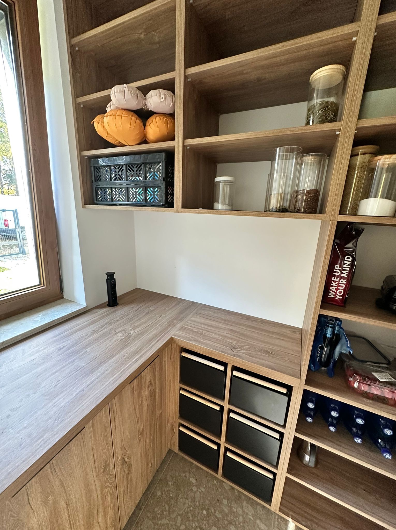 Drewniana zabudowa kuchenna z półkami i szufladami, widok z bliska. Na półkach pojemniki, koszyk i dekoracje. Blat roboczy z gniazdkiem elektrycznym.