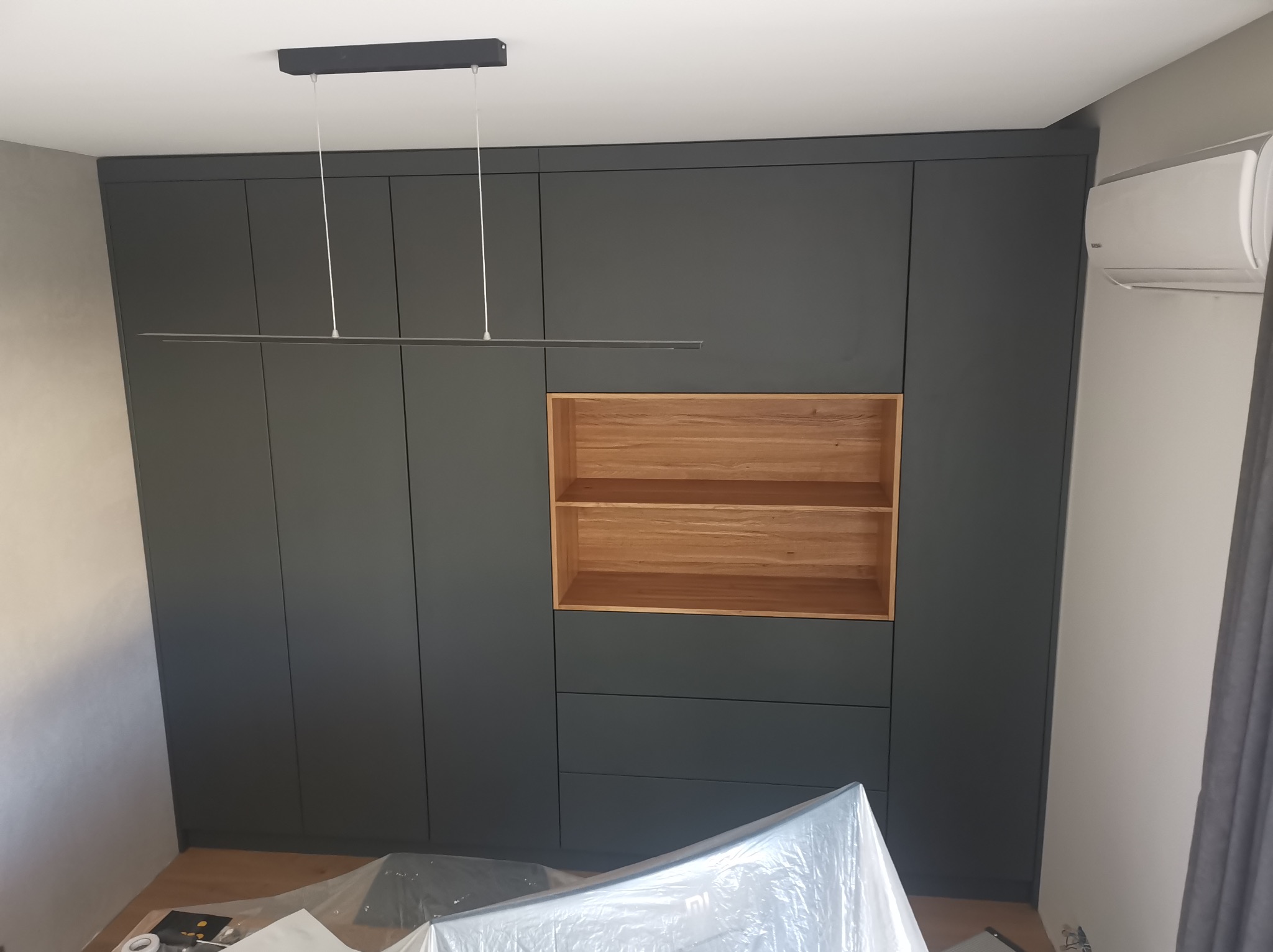 Ciemnoszara szafa wnękowa z drewnianą wstawką i oświetleniem LED, klimatyzator na ścianie. Widoczne zabezpieczenia podłogi folią malarską.
