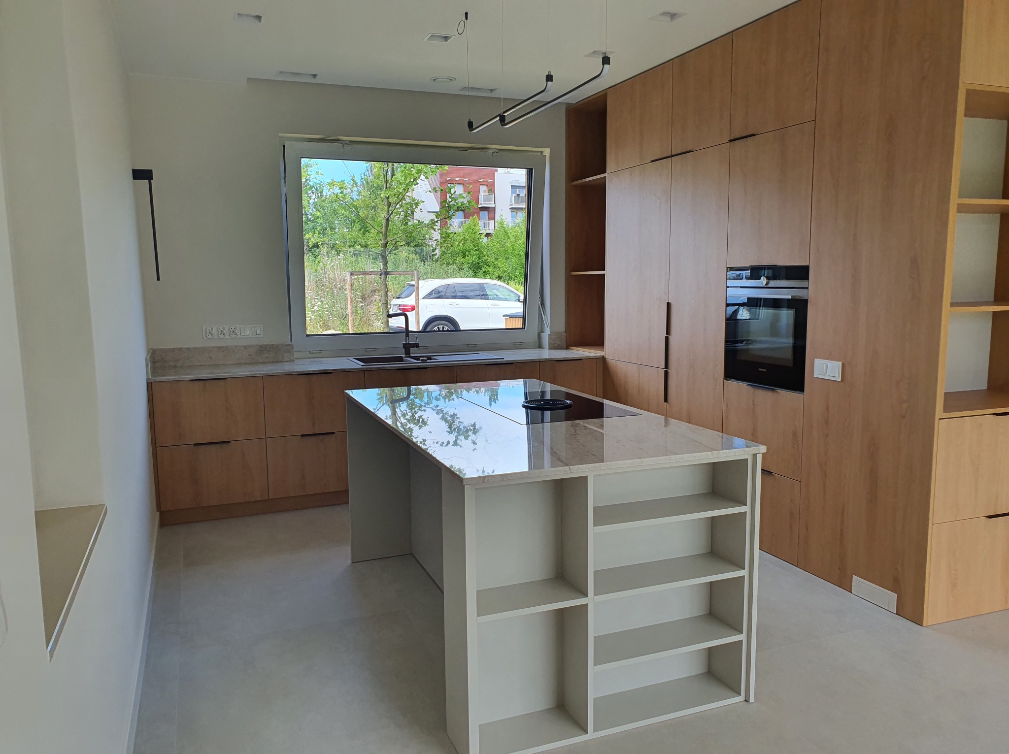 Nowoczesna kuchnia z drewnianymi frontami, wyspą z półkami i blatem z kamienia. W tle okno z widokiem na zieleń i budynek mieszkalny. Minimalistyczne oświetlenie.