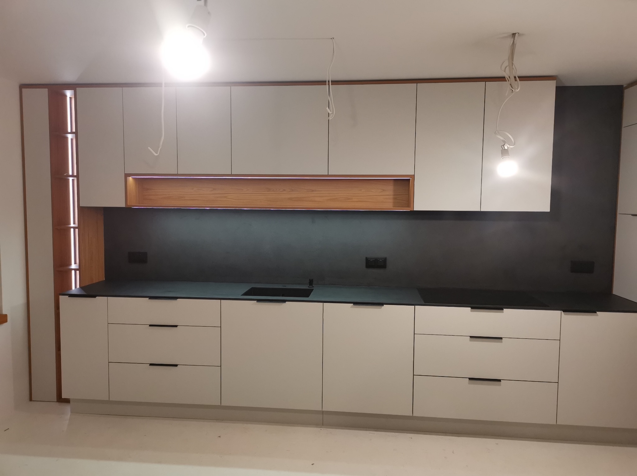 Nowoczesna kuchnia z białymi frontami i czarnym blatem. Górne szafki z drewnianym akcentem i oświetleniem LED. Minimalistyczne uchwyty i gładkie powierzchnie.