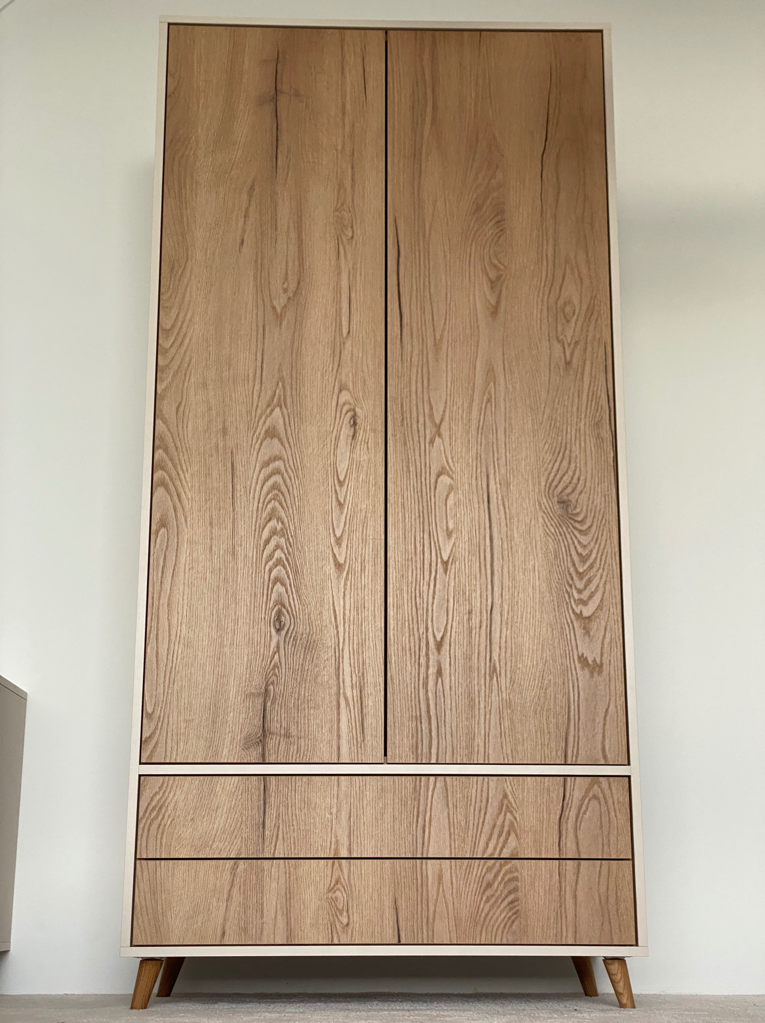 Dwudrzwiowa szafa z trzema szufladami, wykończona okleiną imitującą jasne drewno, na niskich nóżkach, z delikatną, kremową ramą, stojąca na tle białej ściany.