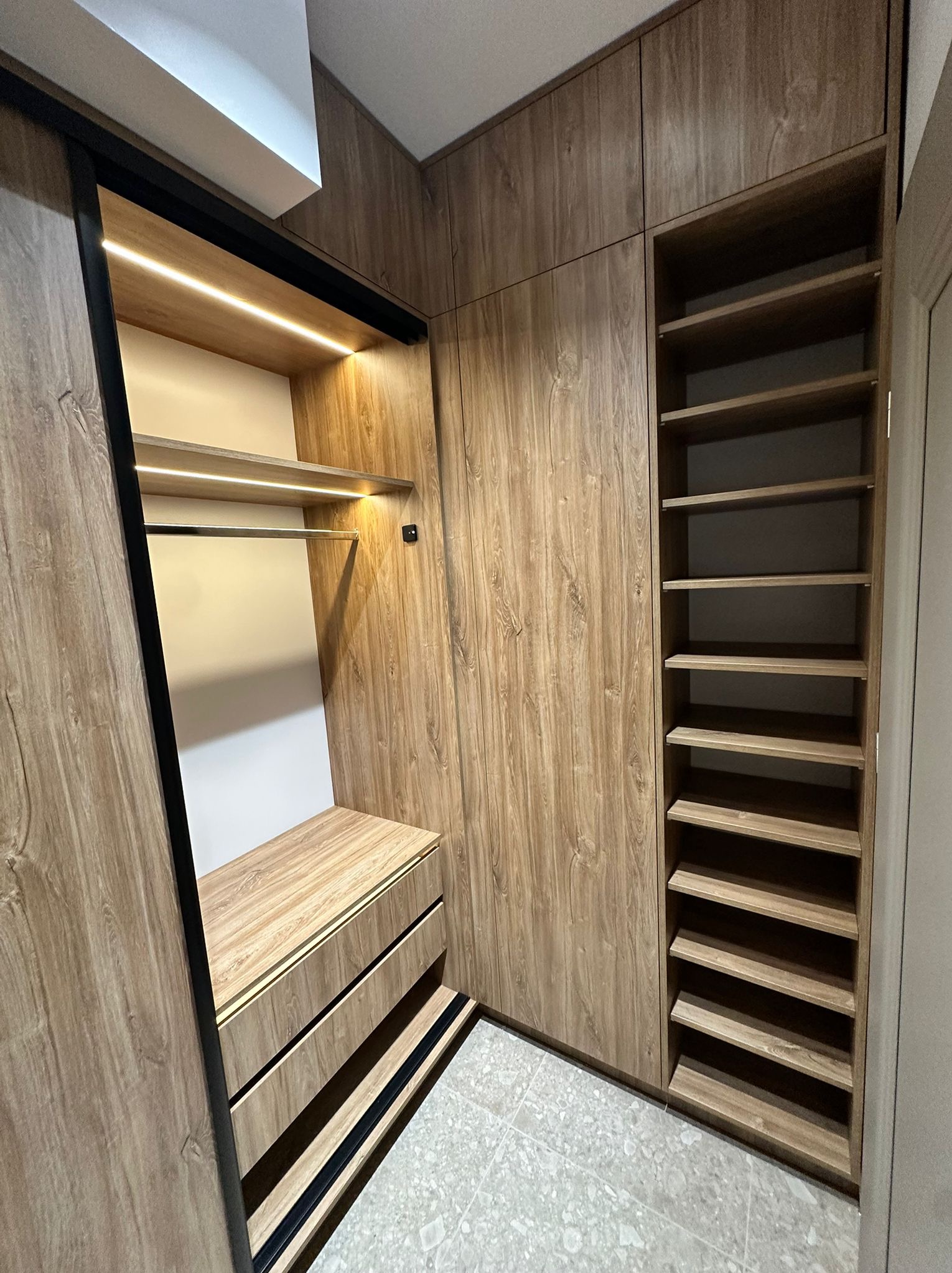 Funkcjonalna szafa wnękowa z przesuwnymi drzwiami i oświetleniem LED, wykończona w ciepłym odcieniu drewna. Widoczne półki i szuflady z czarnymi akcentami.