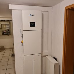 ENOX Robert Koprek - Biały magazyn energii Q CELLS zamontowany na ścianie w pomieszczeniu. Widoczna instalacja elektryczna i fragment drzwi. Minimalistyczny design.