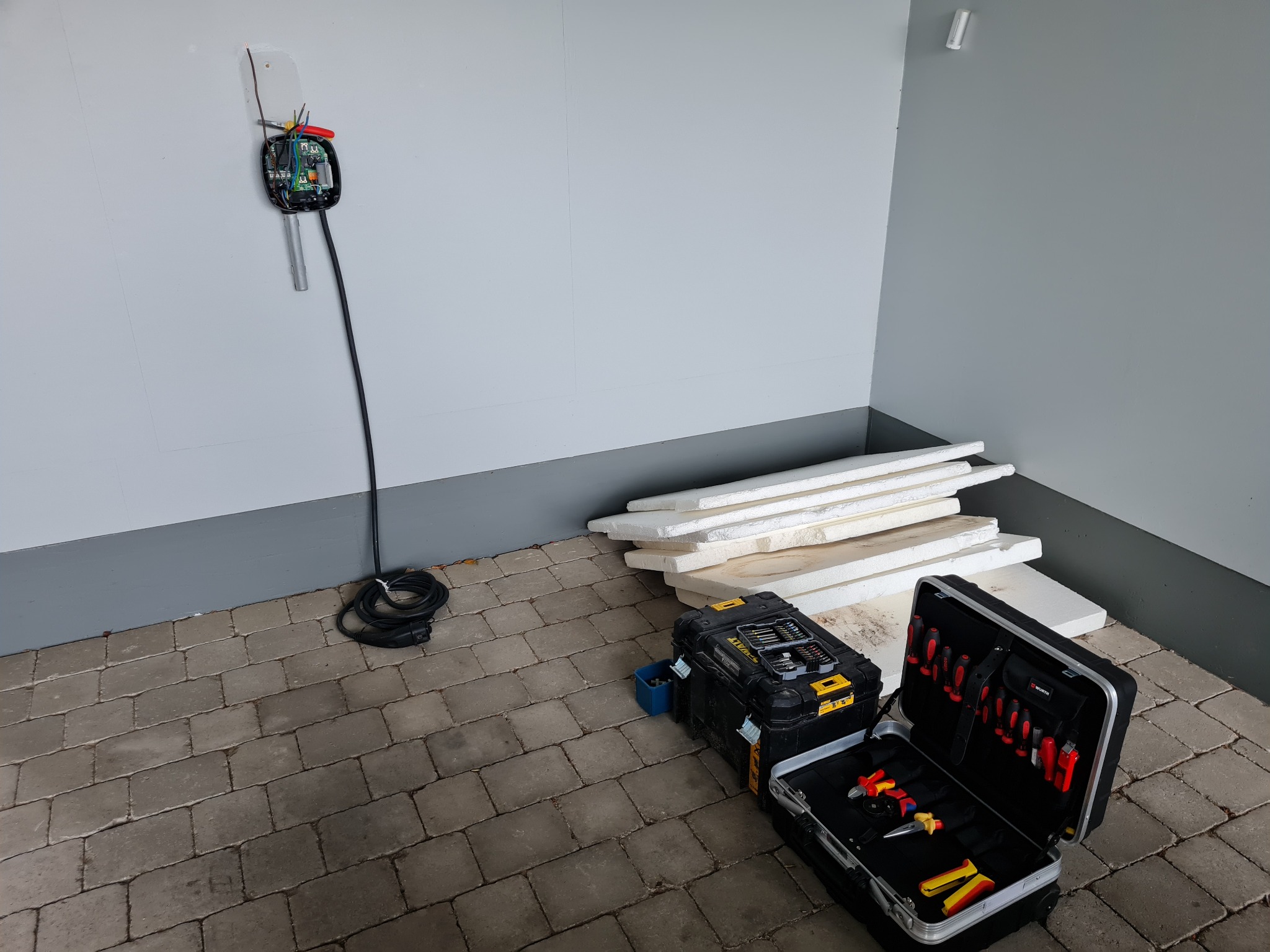 Niedokończona instalacja ładowarki EV na ścianie garażu, obok skrzynki z narzędziami i izolacji. Widoczne przewody i elementy elektryczne.