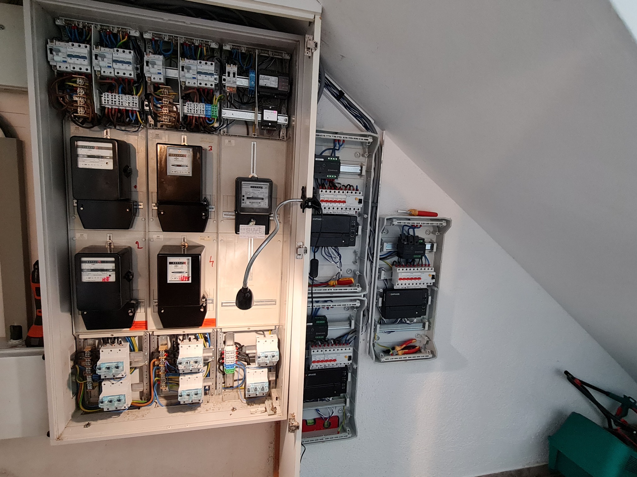 Otwarta skrzynka elektryczna z licznikami i bezpiecznikami, dodatkowa mniejsza skrzynka obok, widoczne narzędzia, instalacja w białym pomieszczeniu ze skośnym sufitem.