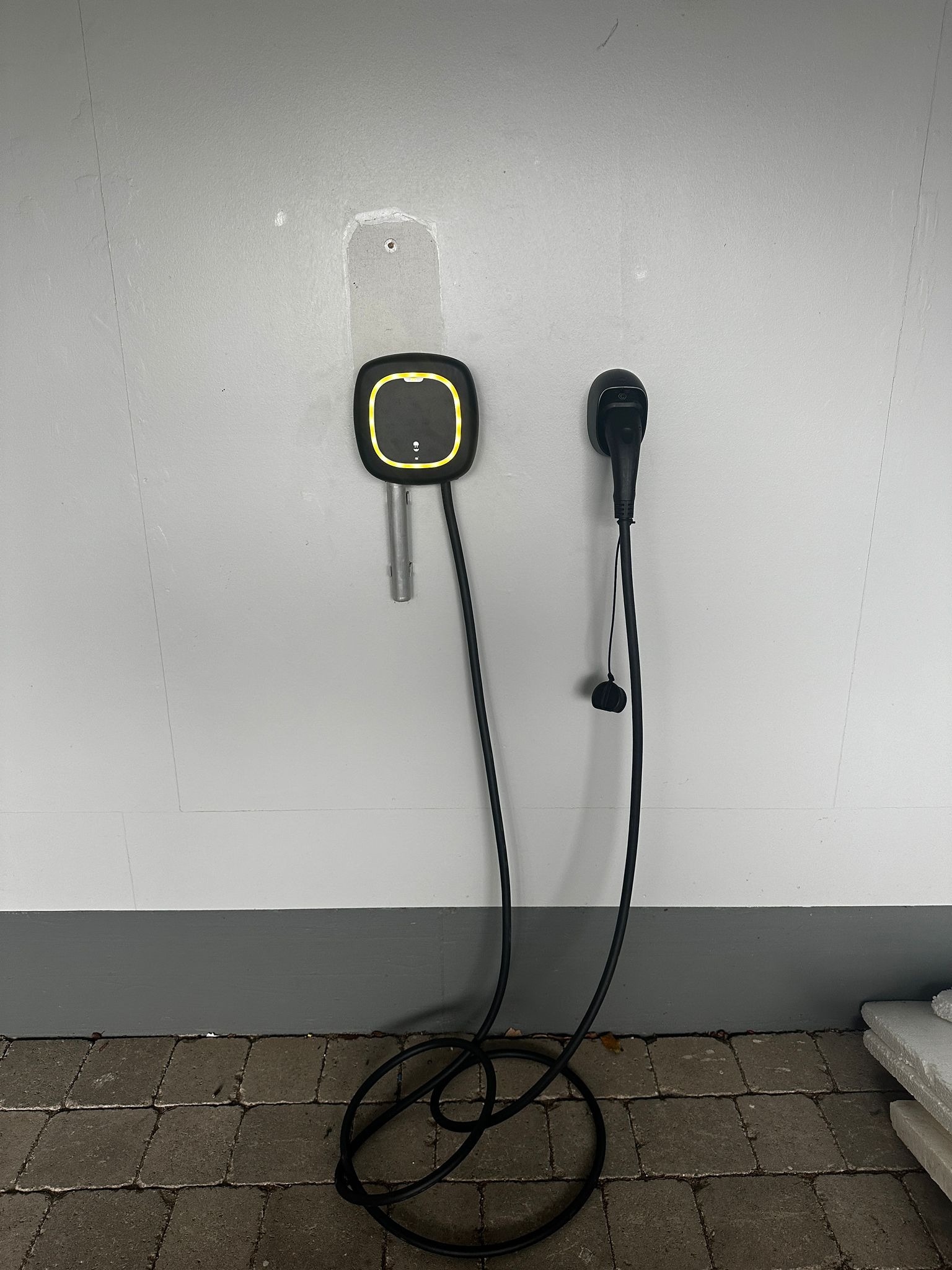 Zamontowana na ścianie ładowarka do pojazdów elektrycznych z kablem zwisającym na podłogę z kostki brukowej. Widoczne ślady po poprzednim montażu.