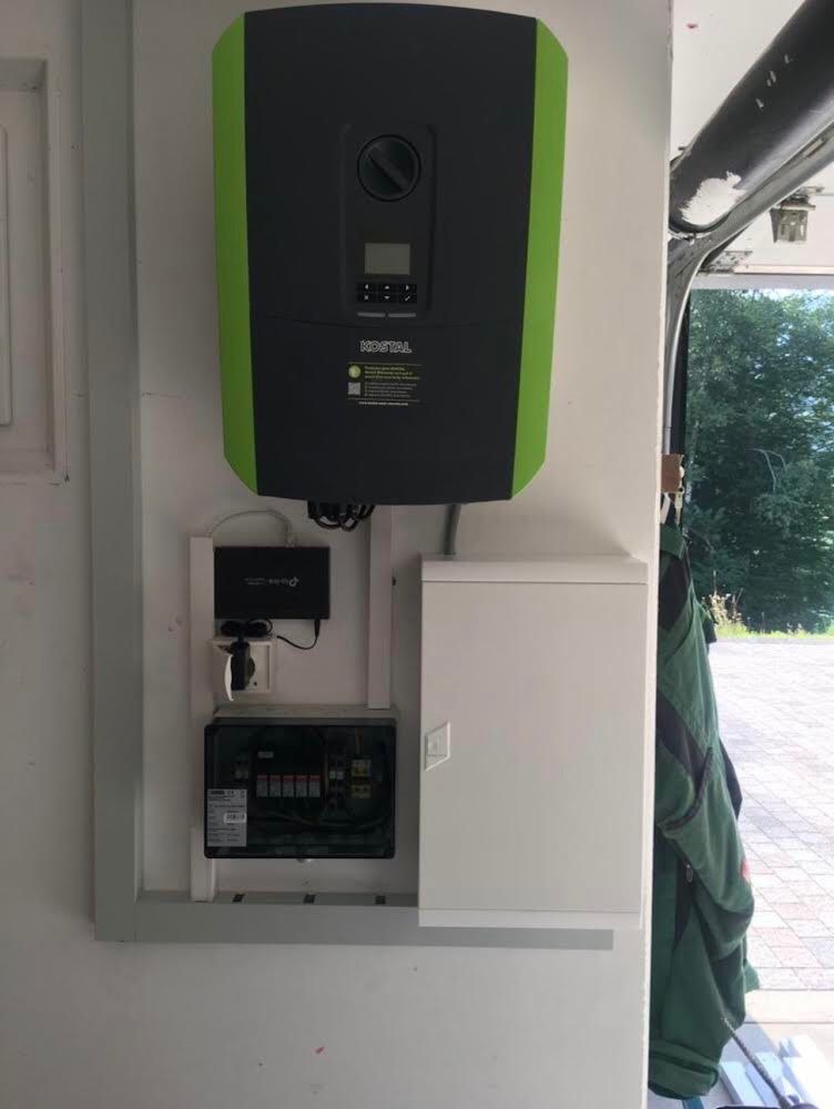 Zamontowany, czarno-zielony magazyn energii Kostal na białej ścianie, obok skrzynka z bezpiecznikami i router. Widoczny fragment otwartej bramy garażowej.