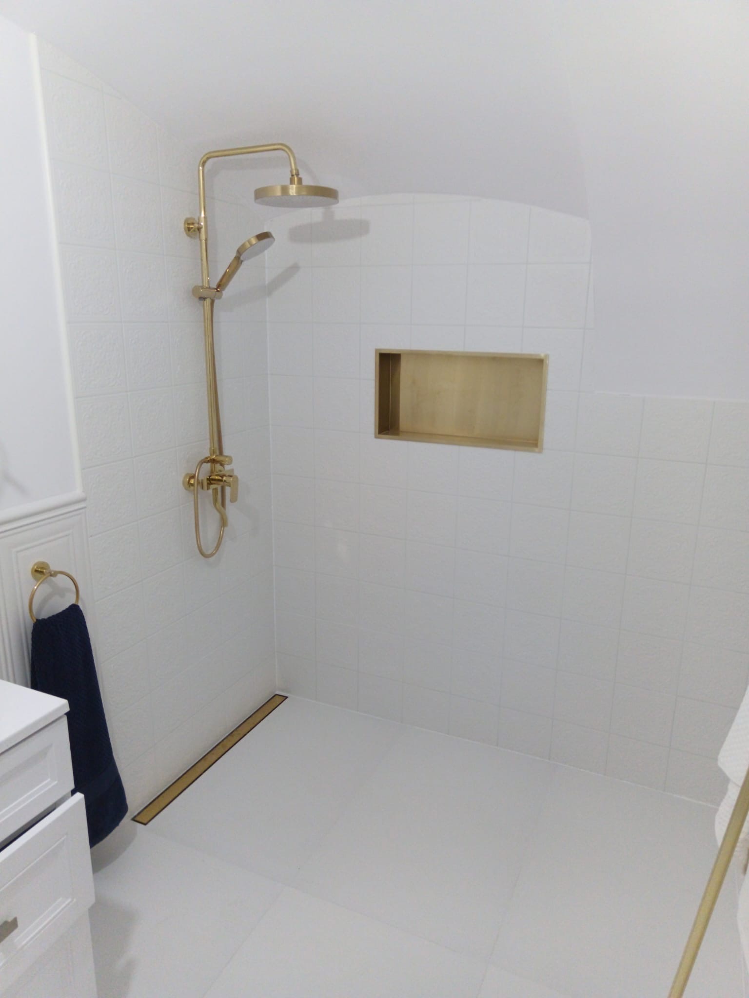 Biała łazienka z kabiną prysznicową typu walk-in, złota armatura, odpływ liniowy i wnęka w ścianie. Minimalistyczny design, jasne płytki z subtelną fakturą.