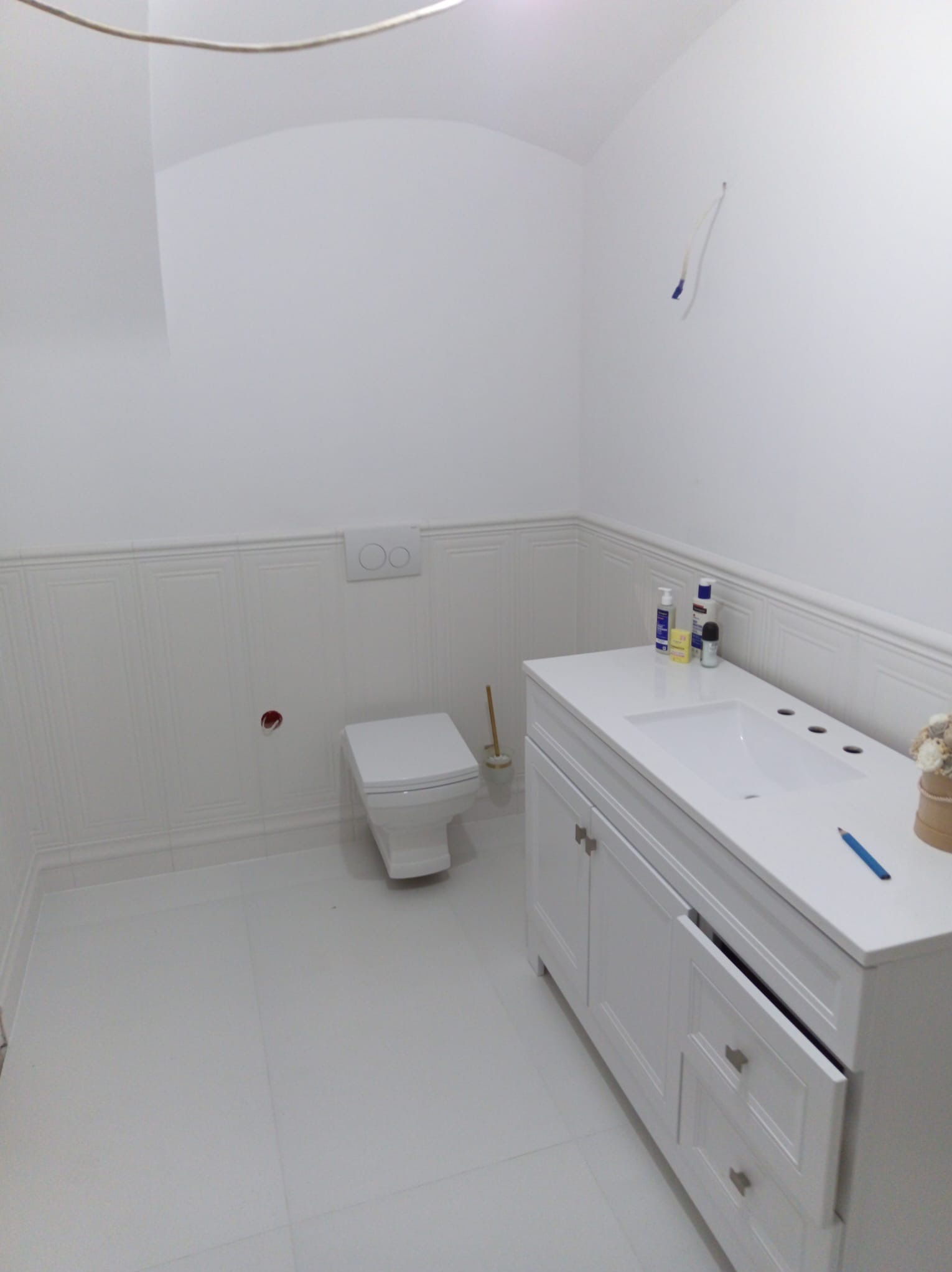 Biała łazienka po remoncie: toaleta, umywalka z szafką, białe płytki imitujące boazerię. Widoczne kosmetyki i ołówek na blacie. Otwarta szuflada szafki.