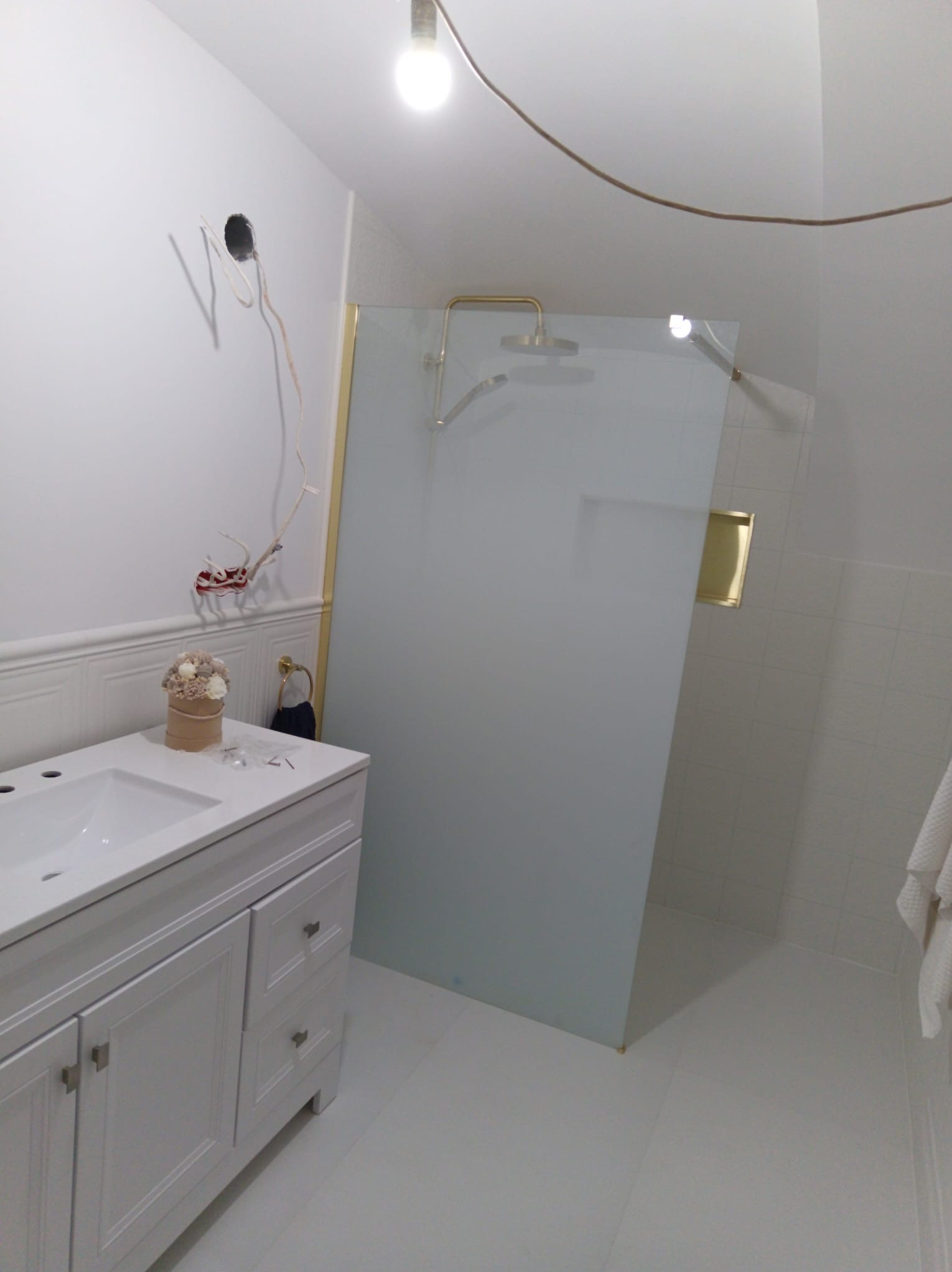 Biała szafka pod umywalkę z dekoracją, kabina prysznicowa ze złotymi akcentami, widoczne przewody elektryczne. Wnętrze łazienki w trakcie wykańczania.