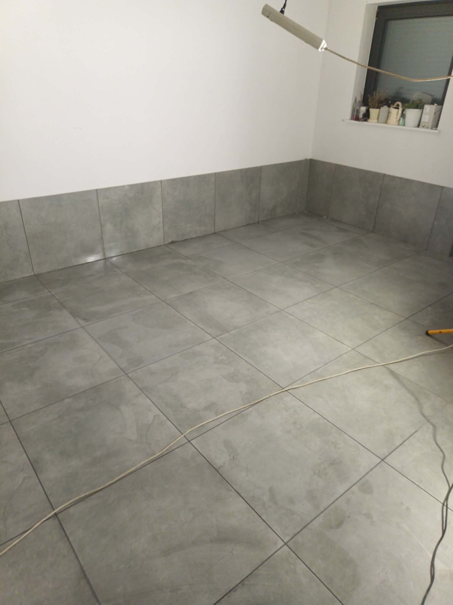 Wykończona podłoga i fragment ściany z szarych płytek w pomieszczeniu w Nysie. Widoczne fugi i kabel na podłodze. Na parapecie okna stoją doniczki z roślinami.