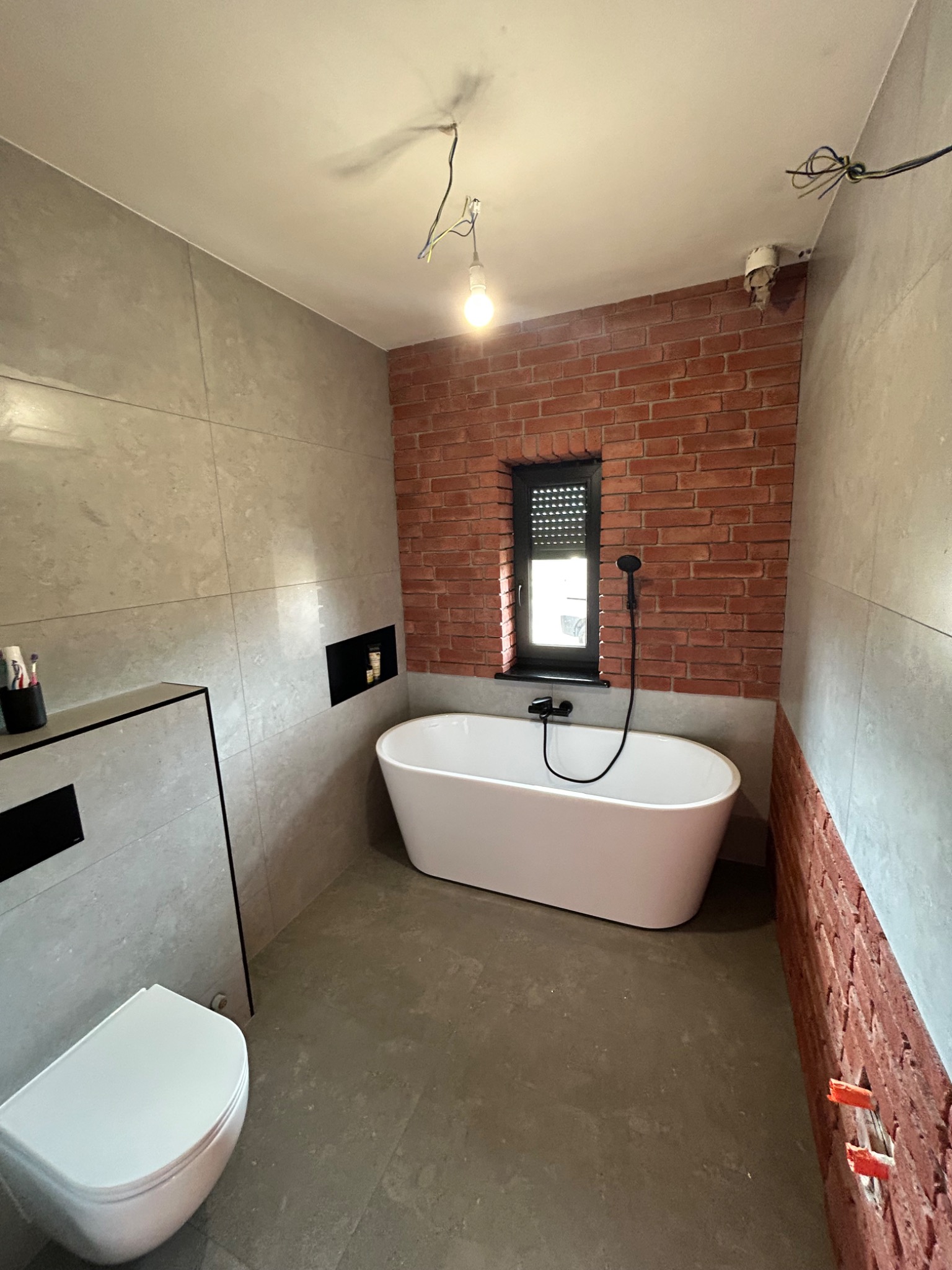 Nowoczesna łazienka w trakcie wykańczania: biała wanna wolnostojąca, szare płytki wielkoformatowe, ściana z czerwonej cegły. Widoczne niedokończone detale.